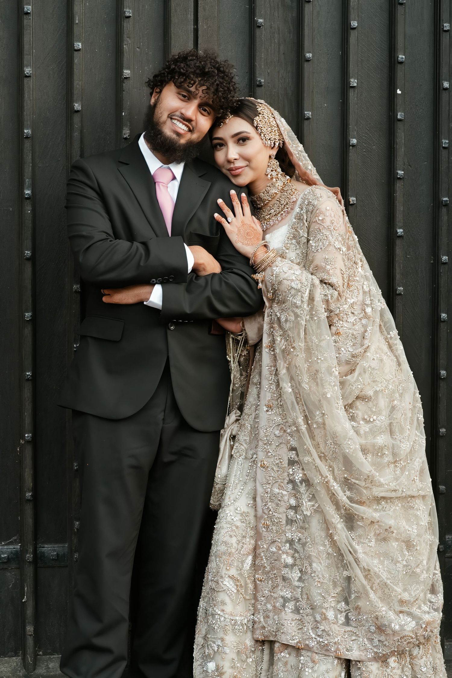 South Asian bride and groom posing together, bride in ornate gold lehenga, groom in black suit with pink tie.