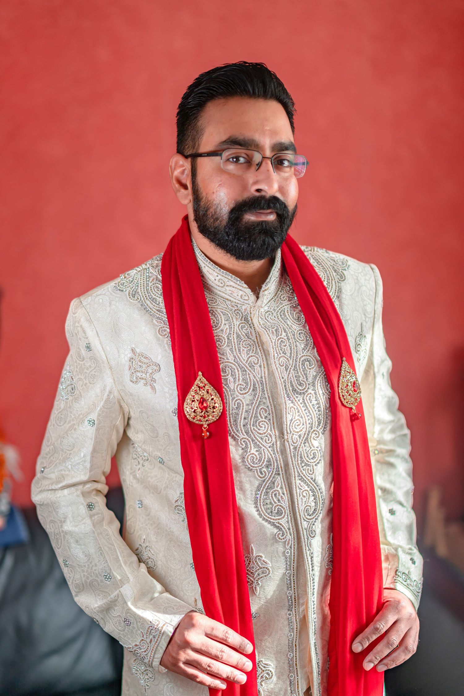 A groom wearing an ornate ivory sherwani with red scarf during a traditional ceremony.