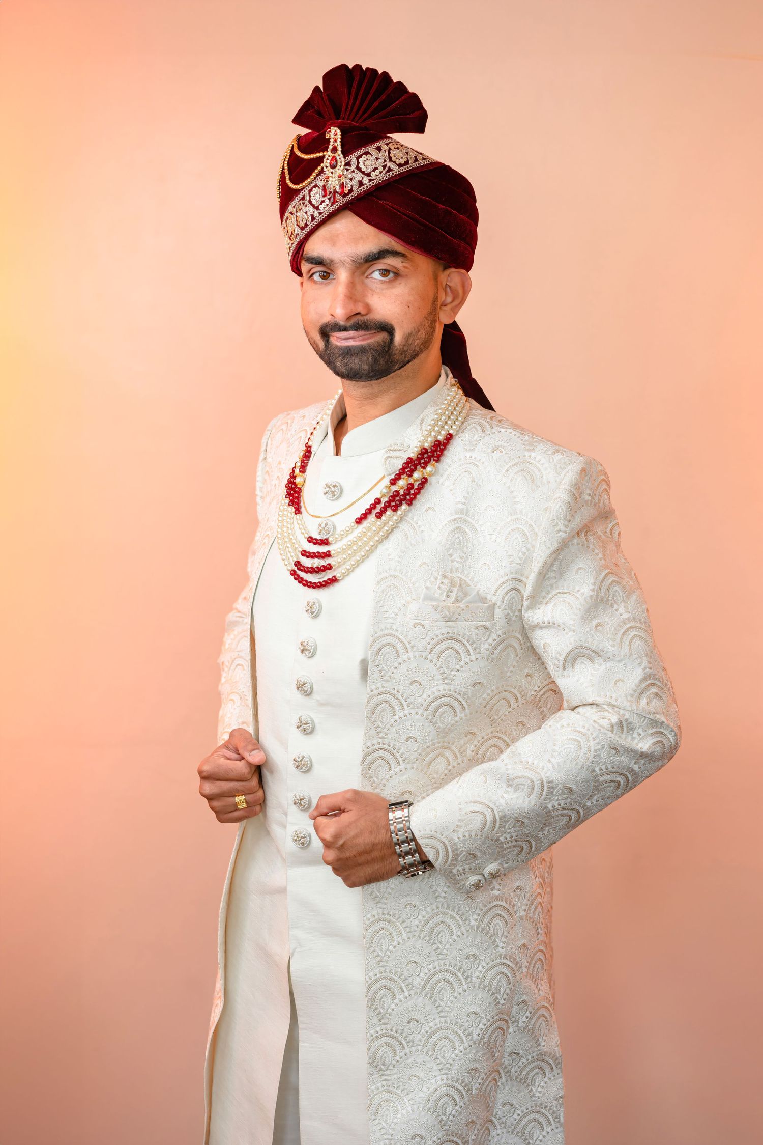 A groom wearing a white sherwani with royal turban and jewelry poses against a peach backdrop.
