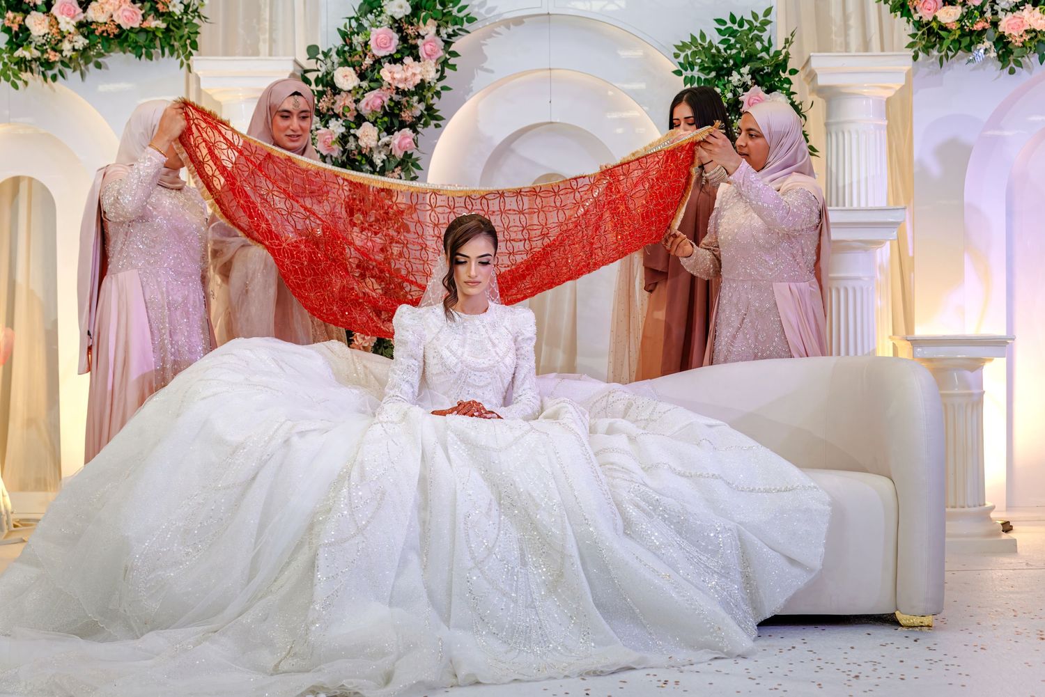 Bride in white gown seated on sofa as attendants hold red lace veil at elegant floral wedding ceremony.