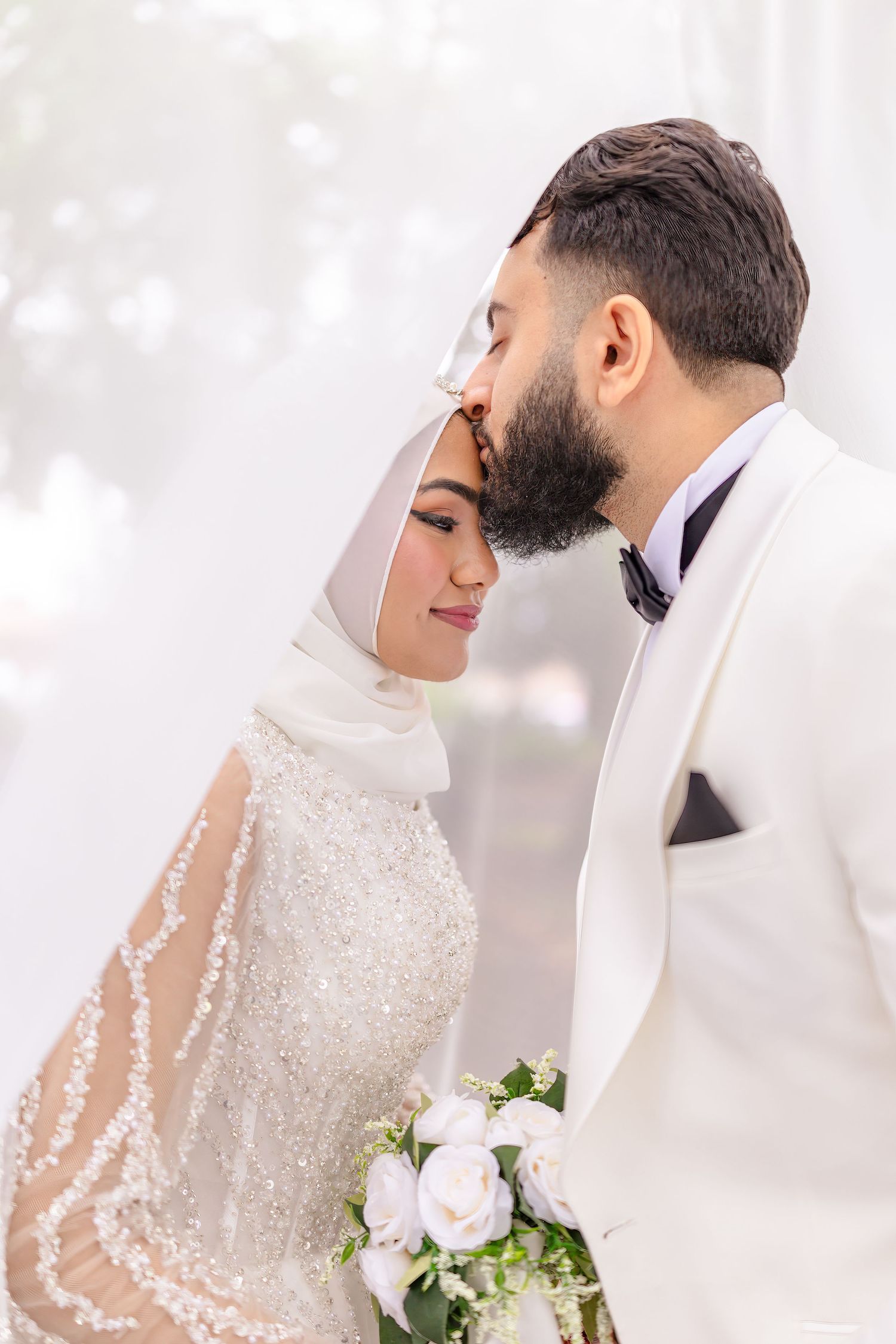 A tender wedding moment captured of a couple in white attire, with the bride in a hijab holding a pink rose bouquet.