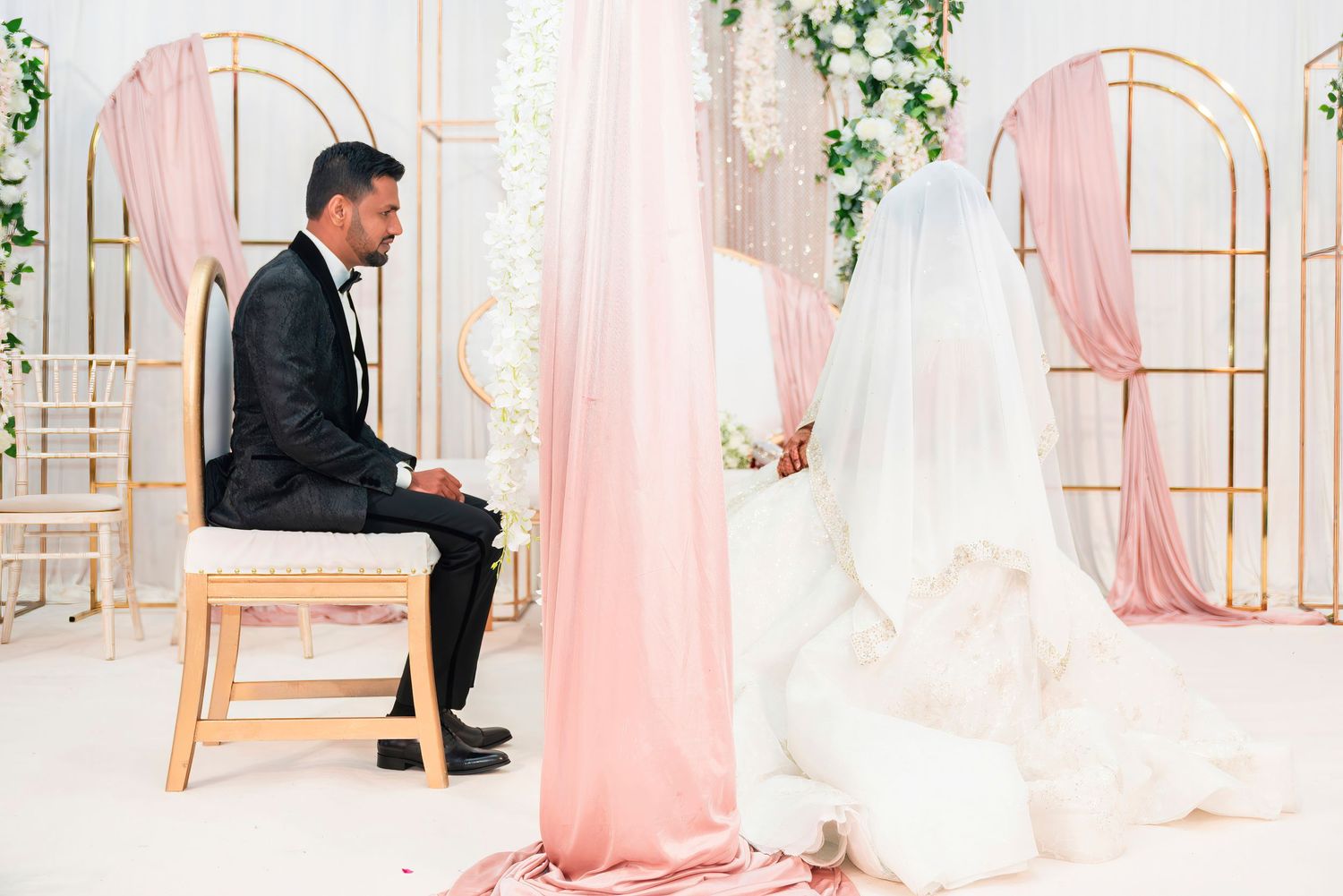 A groom in black suit sits on a wooden chair against a pink backdrop with elegant wedding decor.