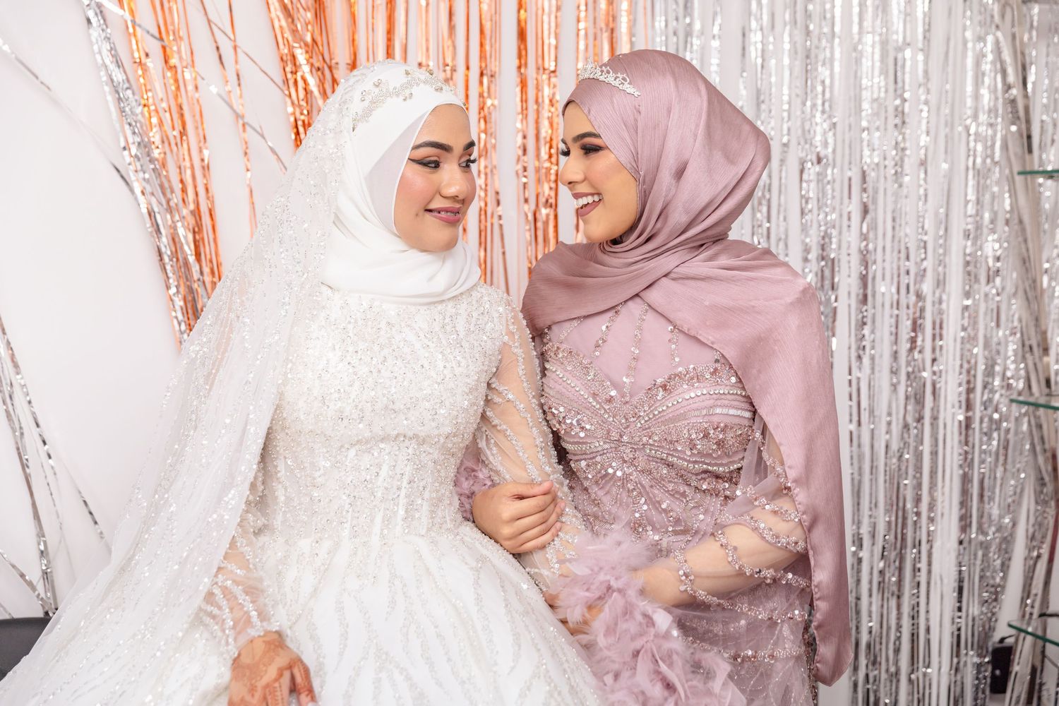 A group of women in hijabs and modest dresses pose together against a silver backdrop at a social gathering.