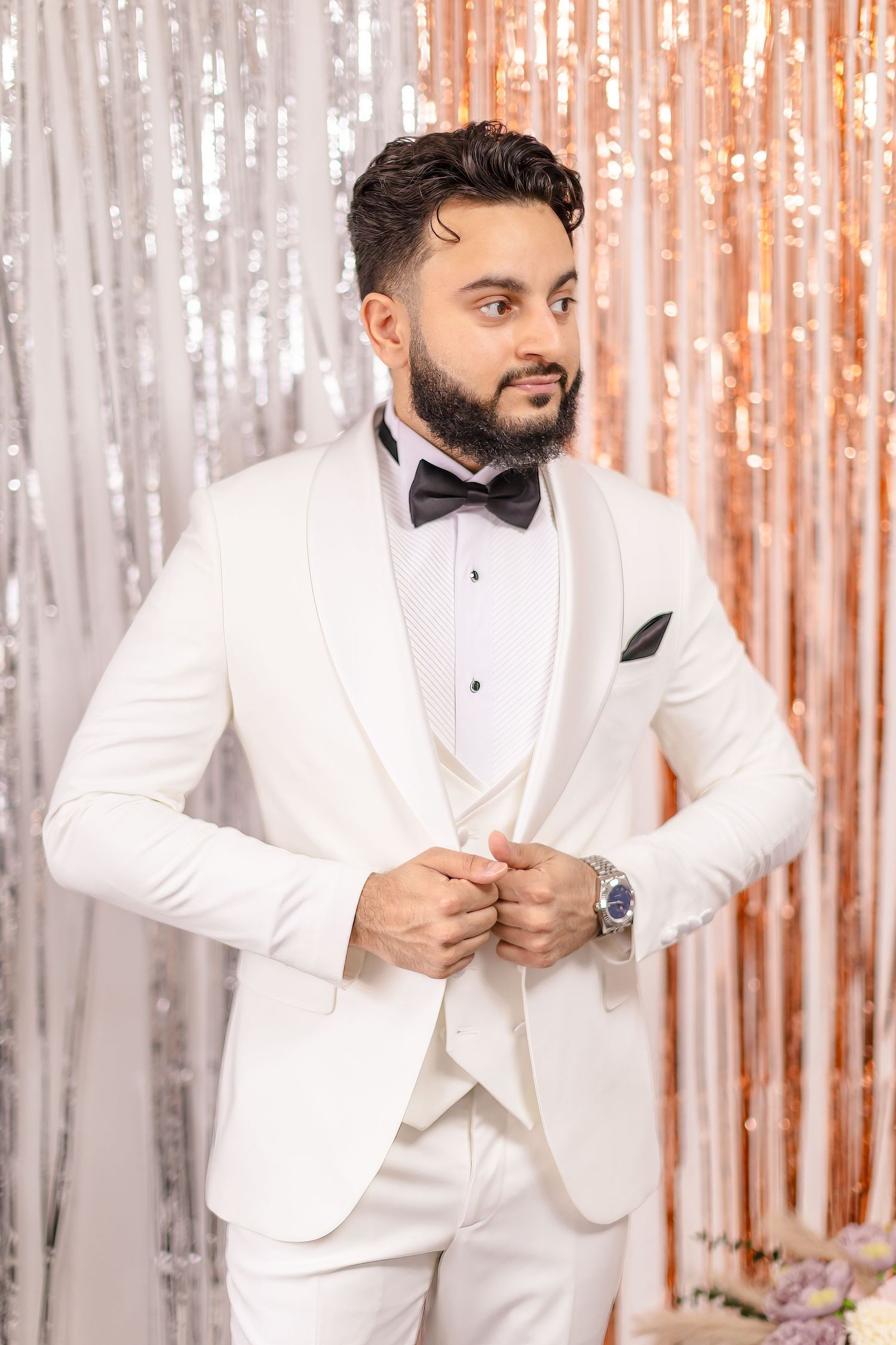 A man wearing a white three-piece tuxedo with black bow tie poses against a shimmery curtain backdrop.