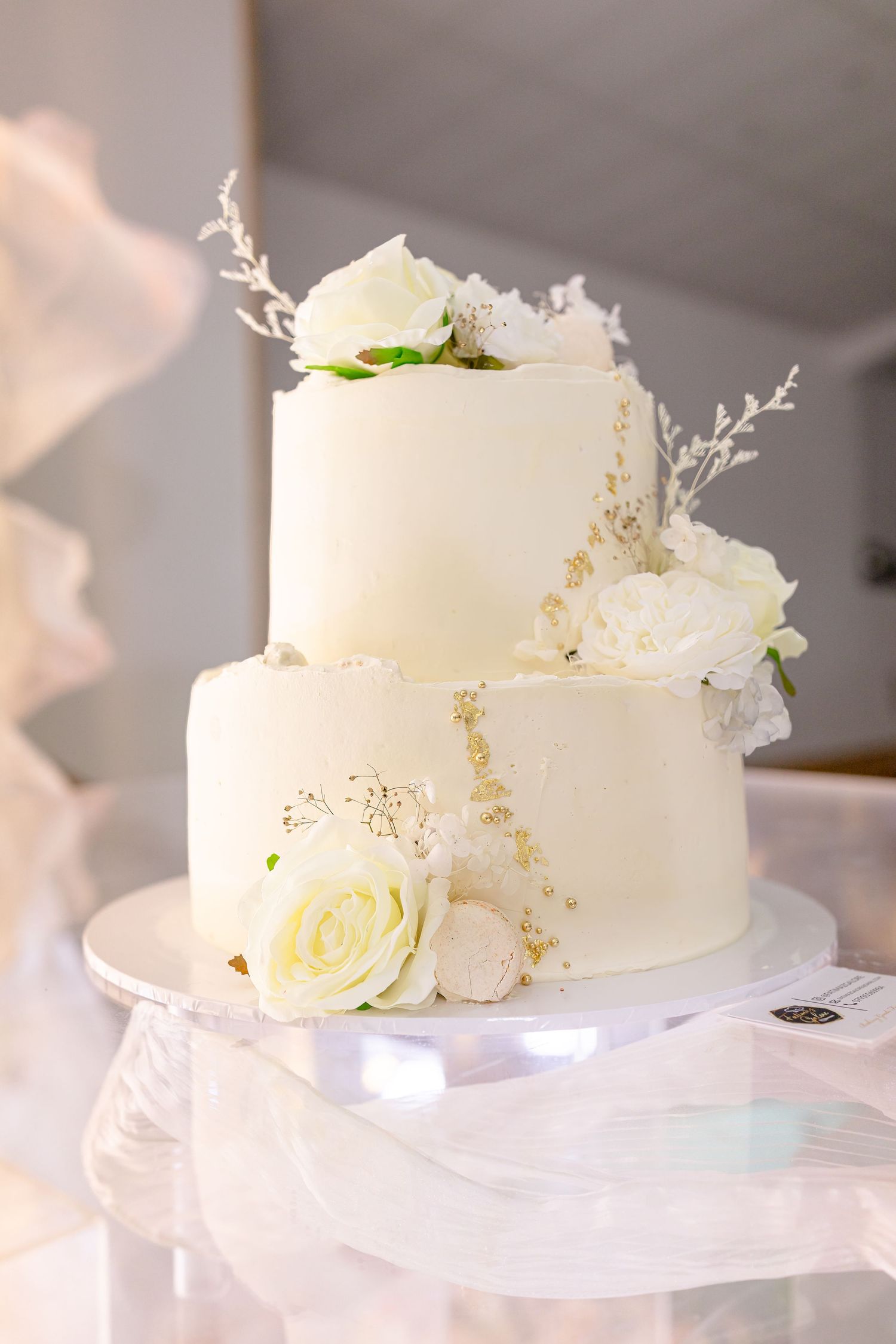 Elegant two-tier white wedding cake decorated with cream roses and delicate white floral accents on a light stand.