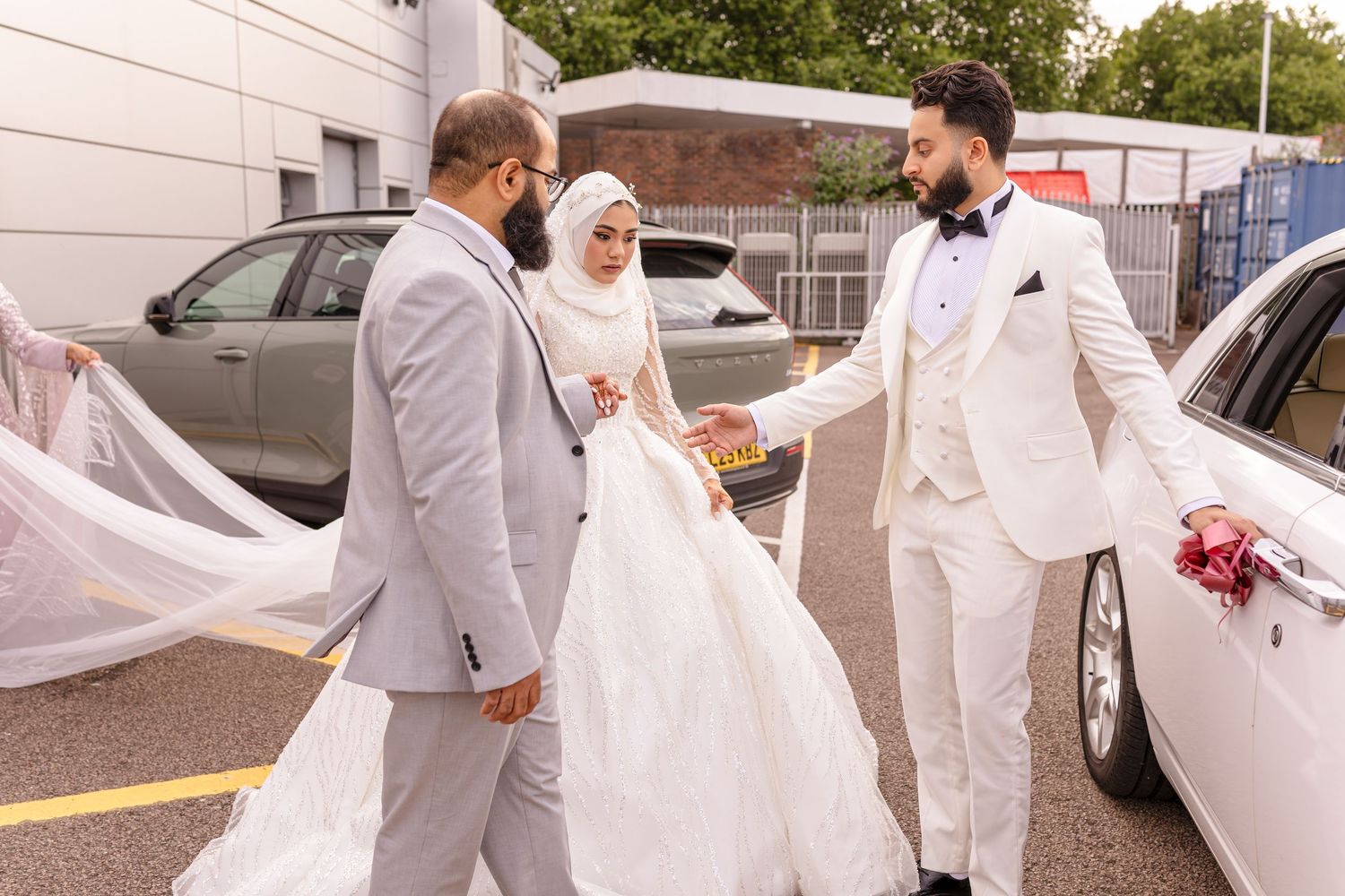 Wedding party in all white and grey formal attire gathered outside near a white car.
