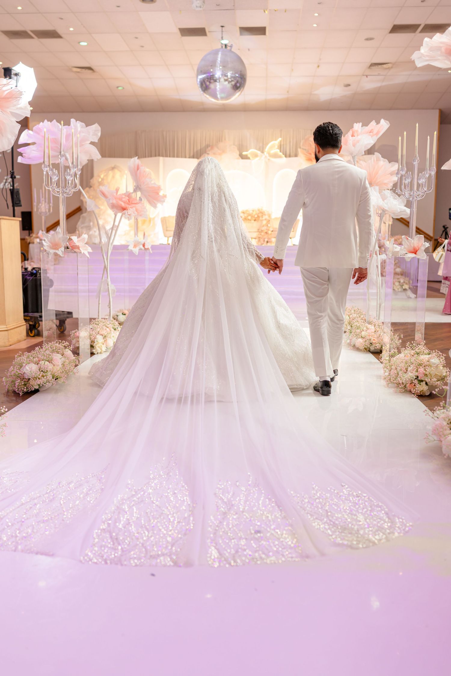 Wedding couple walking down an elegant reception hall with a flowing cathedral veil and white decor.