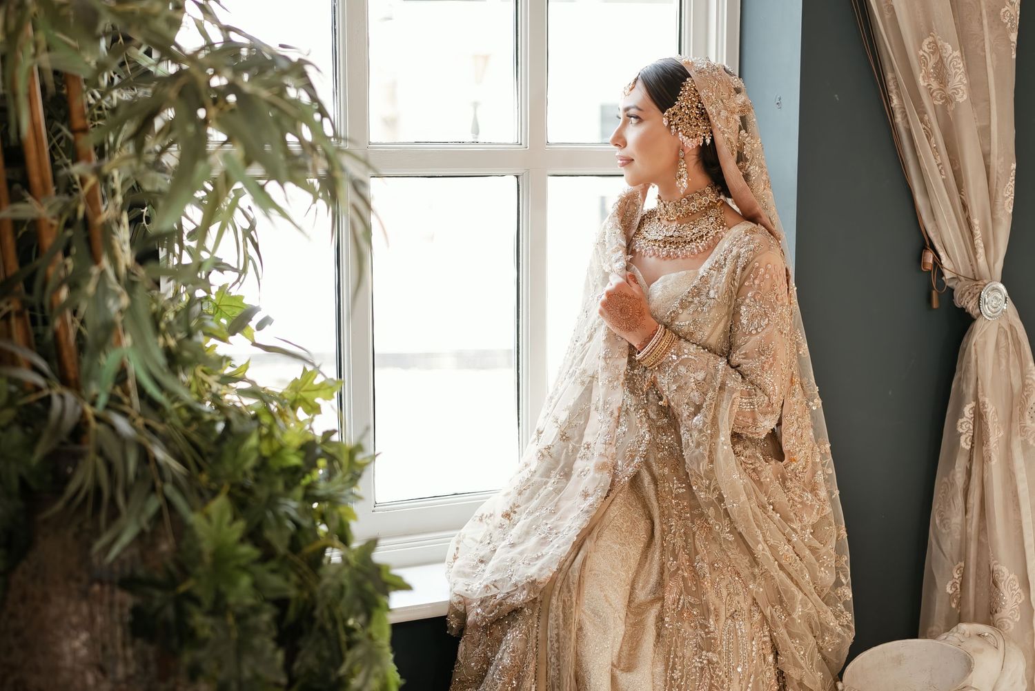 A bride in an ornate gold and ivory South Asian bridal lehenga stands by a window in soft natural light.