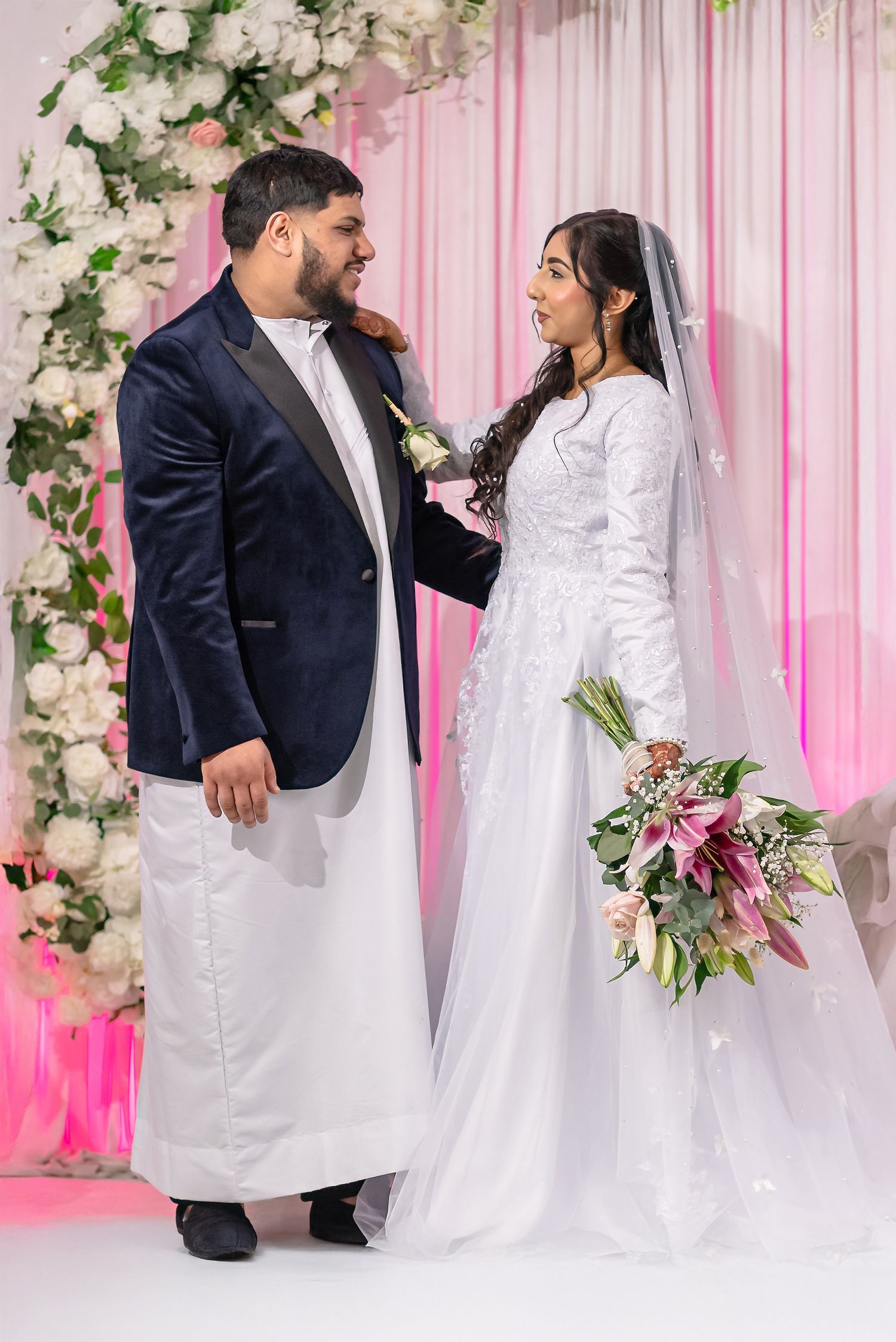A couple celebrates their wedding under a floral arch with pink drapery and white roses.