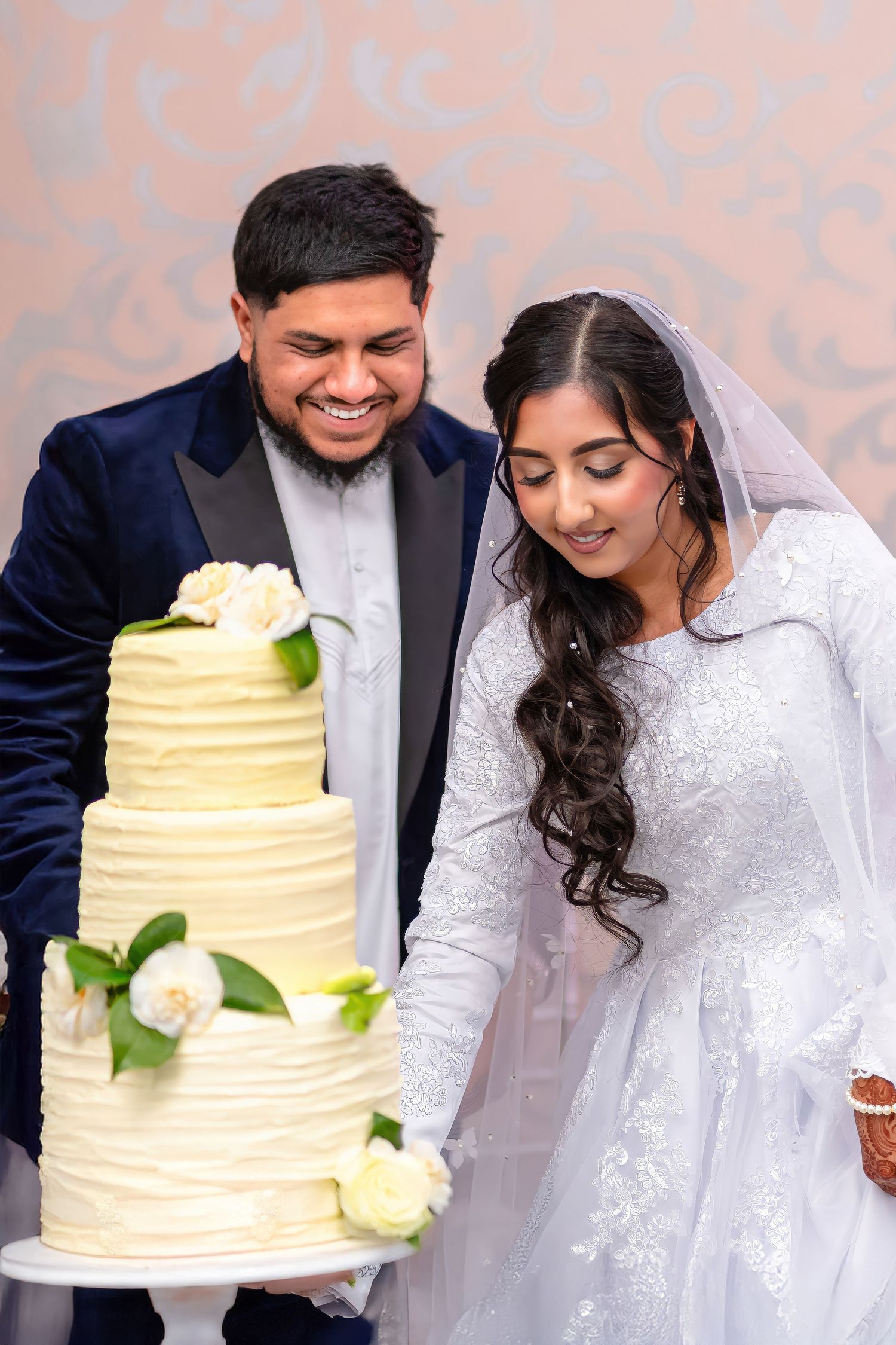 A newlywed couple smiles while cutting their elegant three-tier white wedding cake.