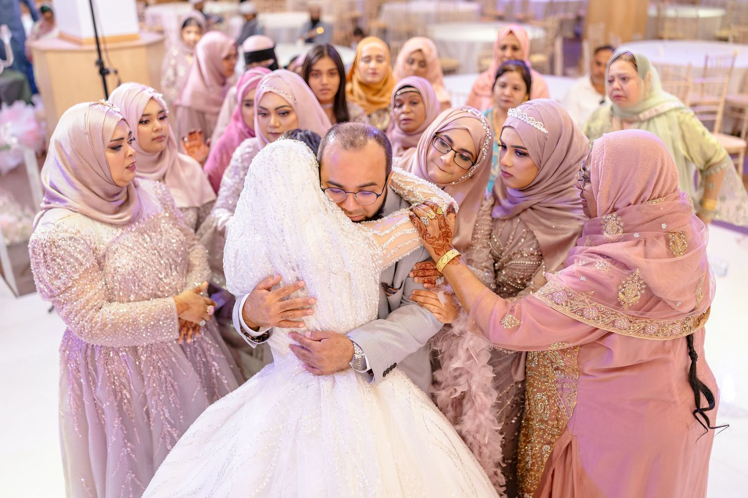 A celebratory wedding scene with guests in pink and purple dresses surrounding the couple at an indoor venue.