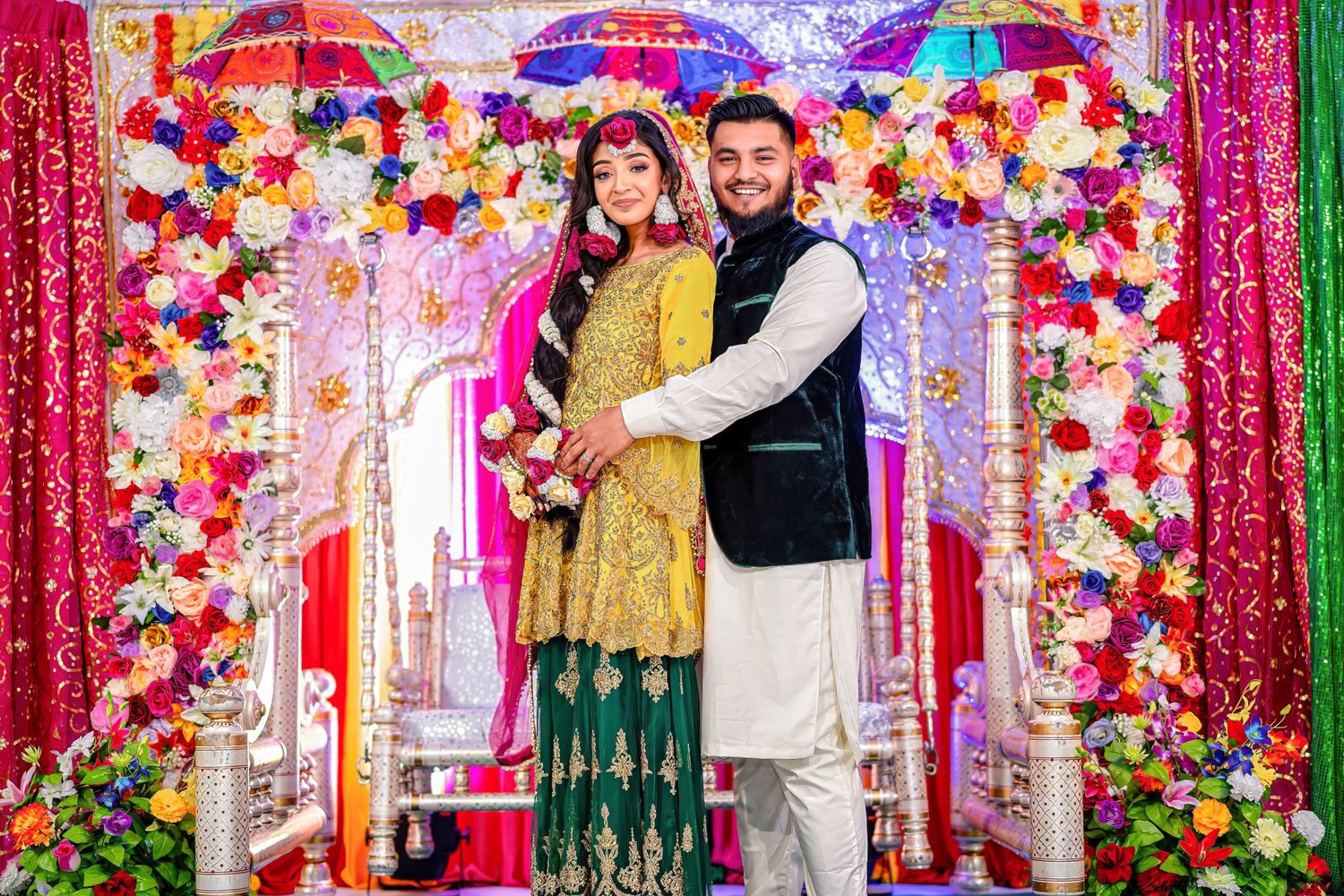 A couple poses under a colorful floral arch with pink drapery and festive decorations at a cultural celebration.