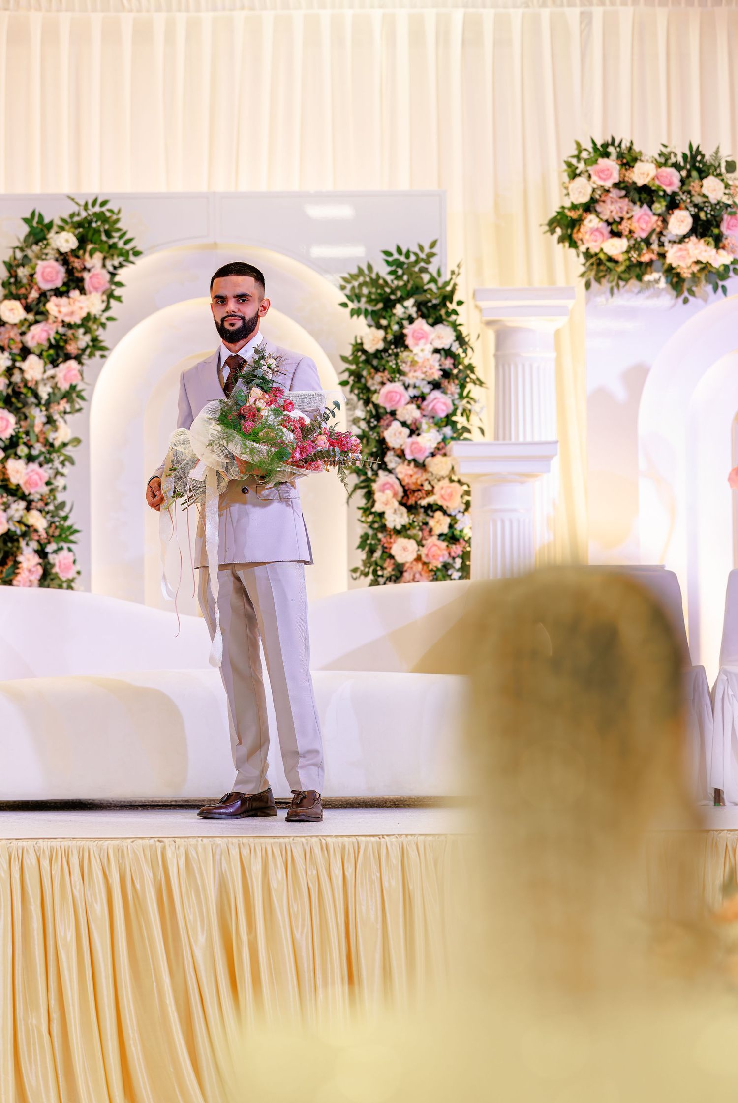 Groom in light gray suit holding bouquet on decorated wedding stage with pink floral arrangements and white columns.