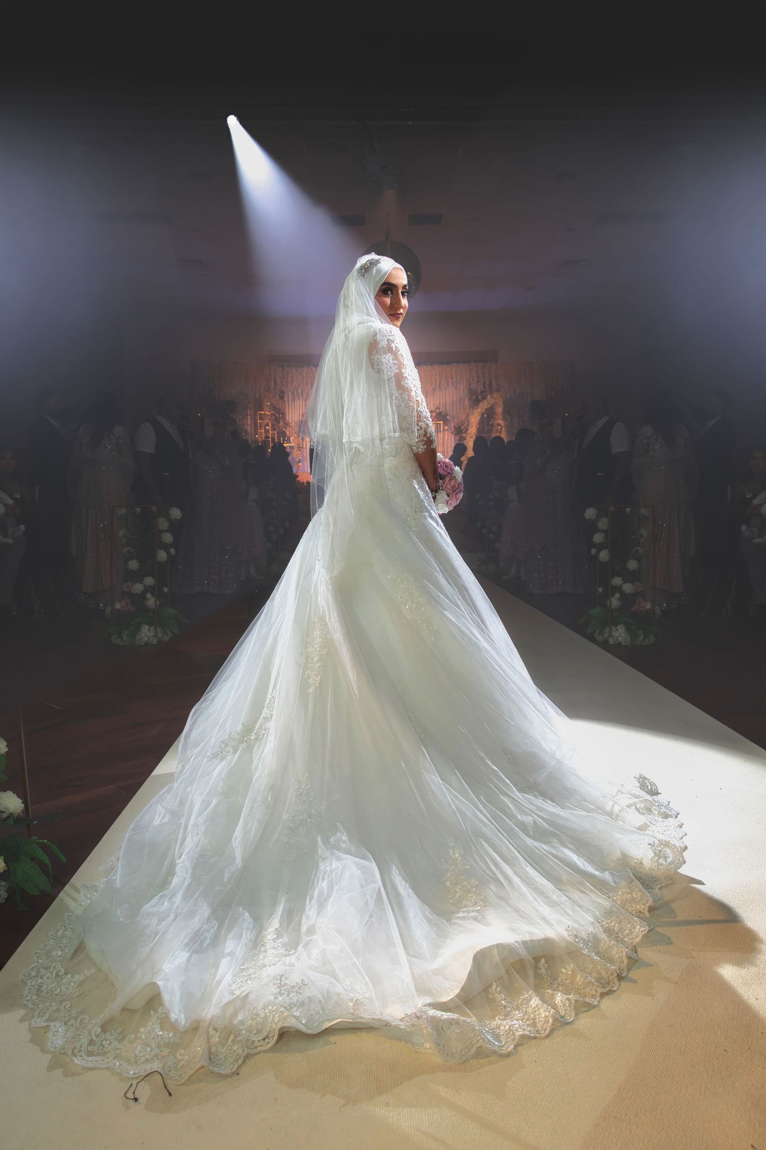 Bride in flowing white gown poses under dramatic spotlight on stage.