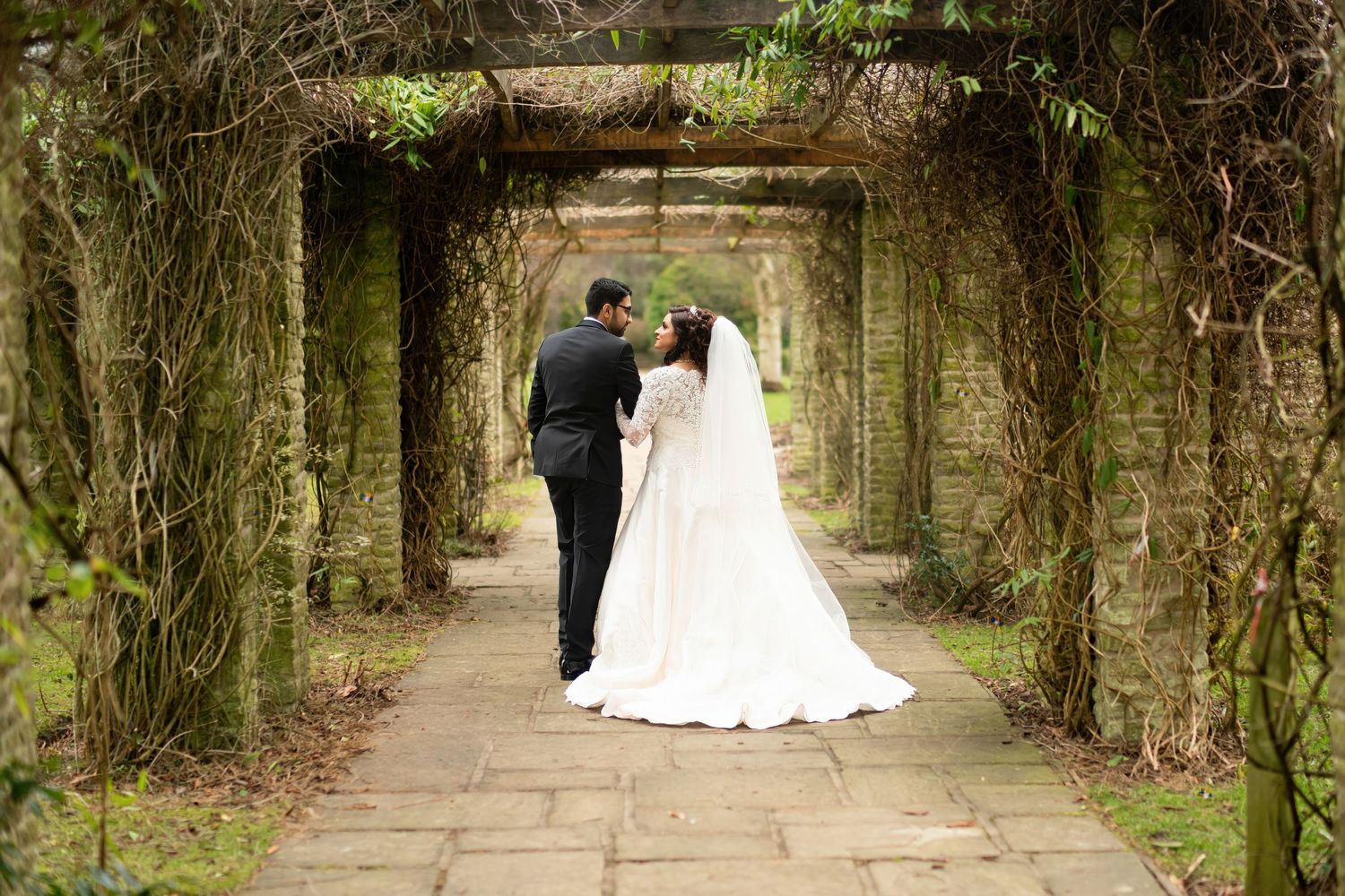 Wedding couple stands beneath vine-covered archway in romantic garden setting.