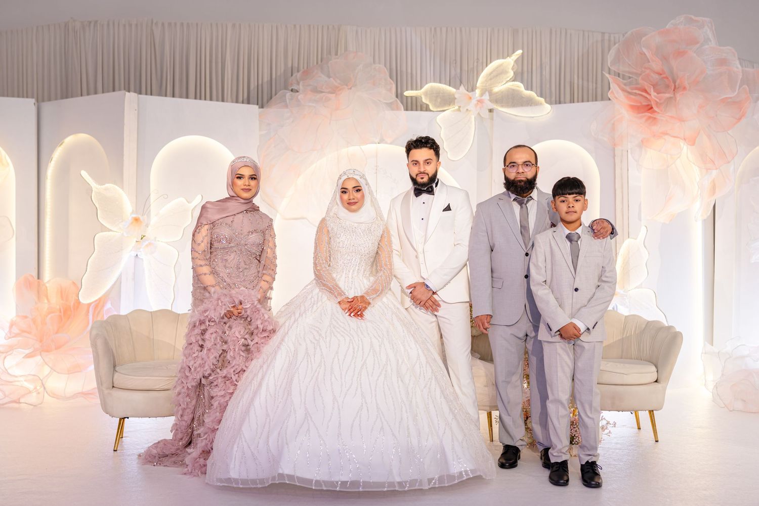 Wedding party poses together against elegant white backdrop with soft pink floral decorations and draped fabric.