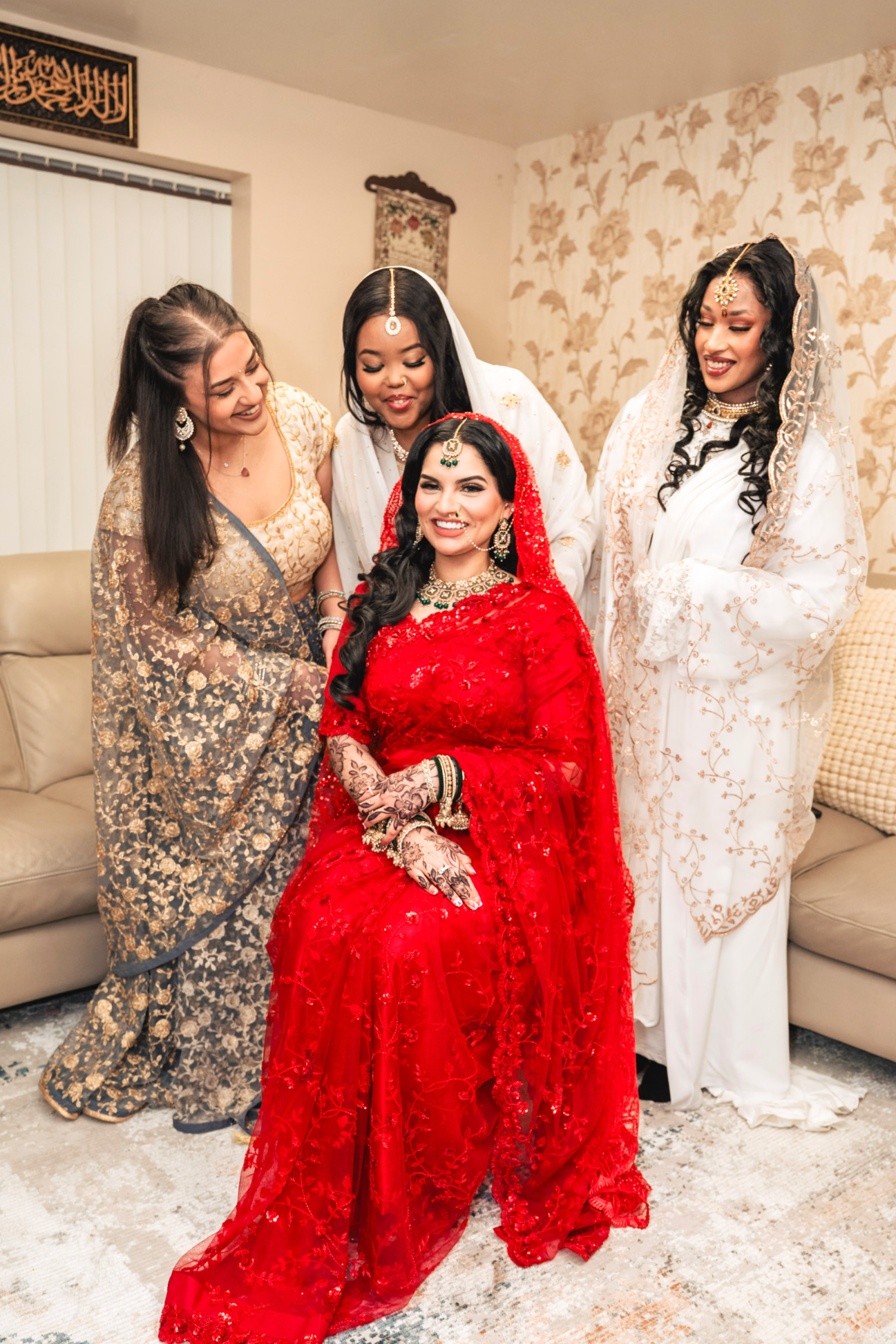 Four women wearing traditional South Asian wedding attire with a bride in red and bridesmaids in sparkly dresses gather indoors.