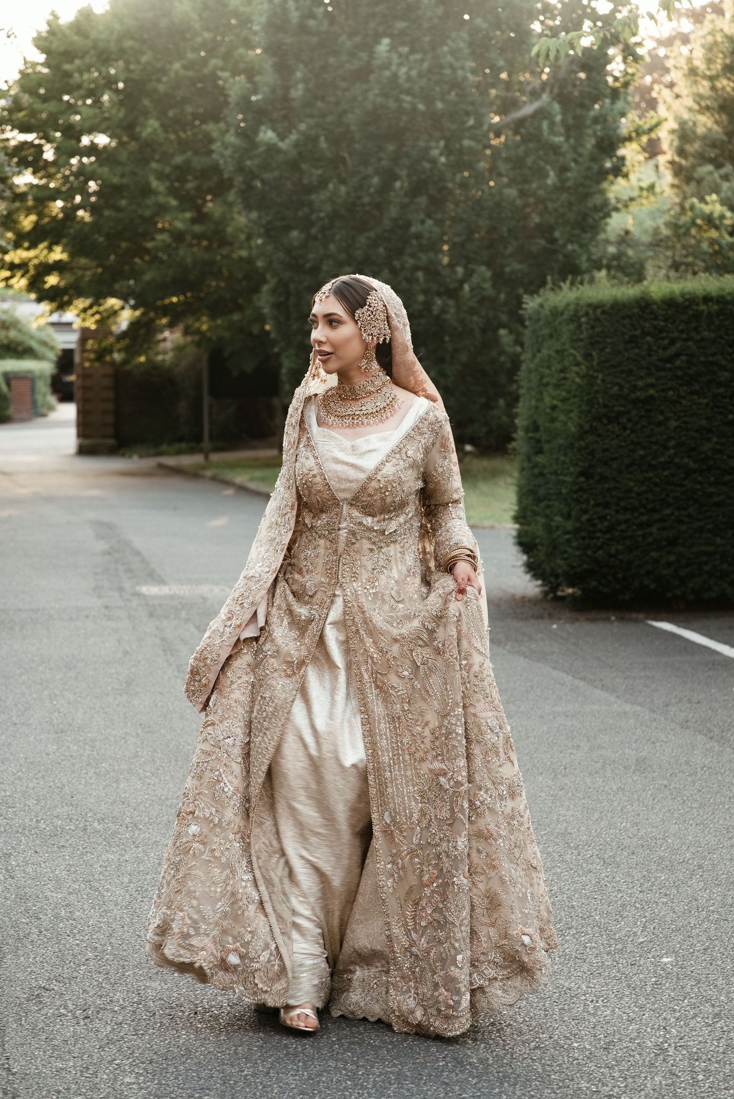 Bride in gold embroidered lehenga and dupatta walks outdoors, showcasing intricate bridal wear.