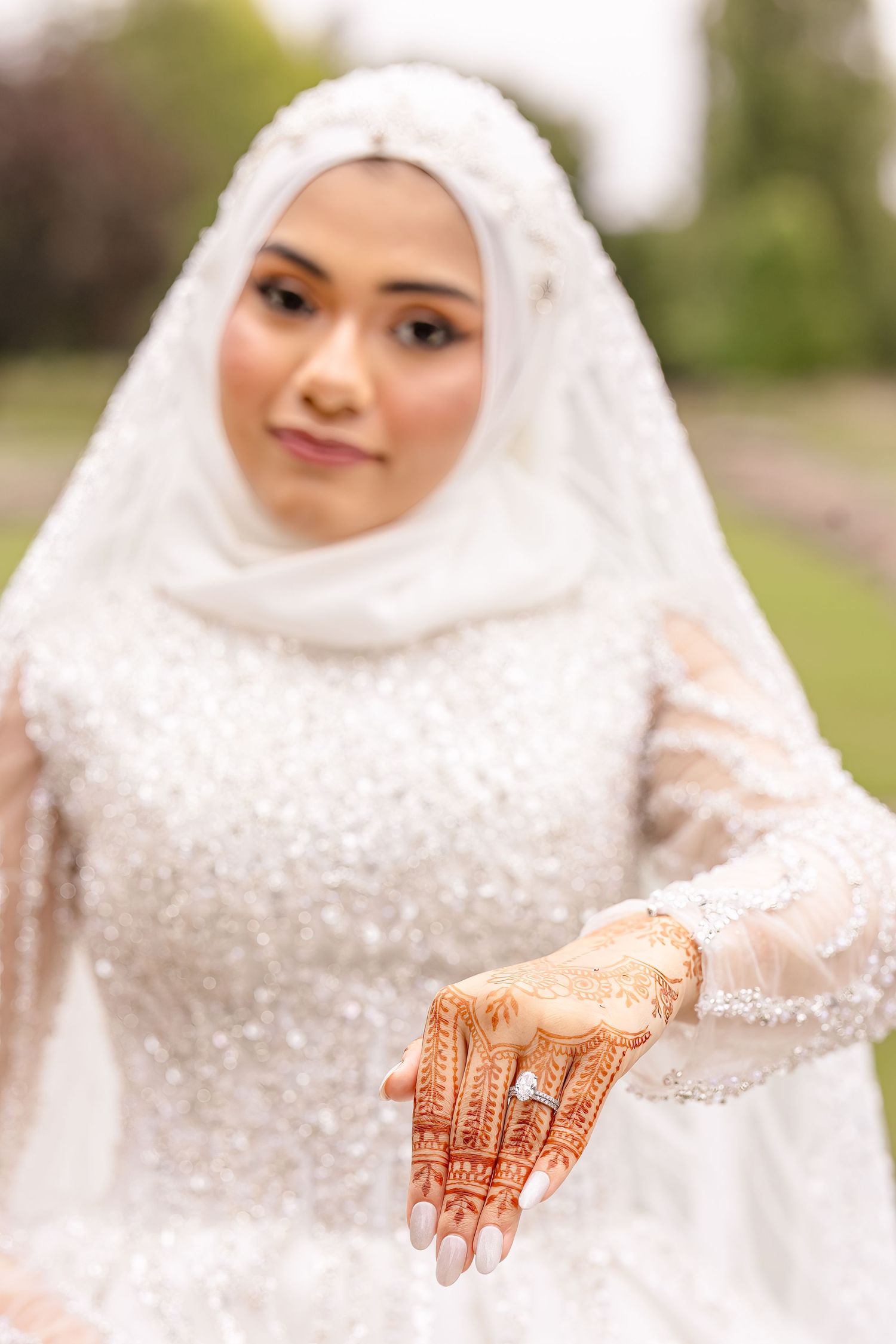 A bride in a white embellished wedding dress and hijab displays traditional henna designs on her hand.