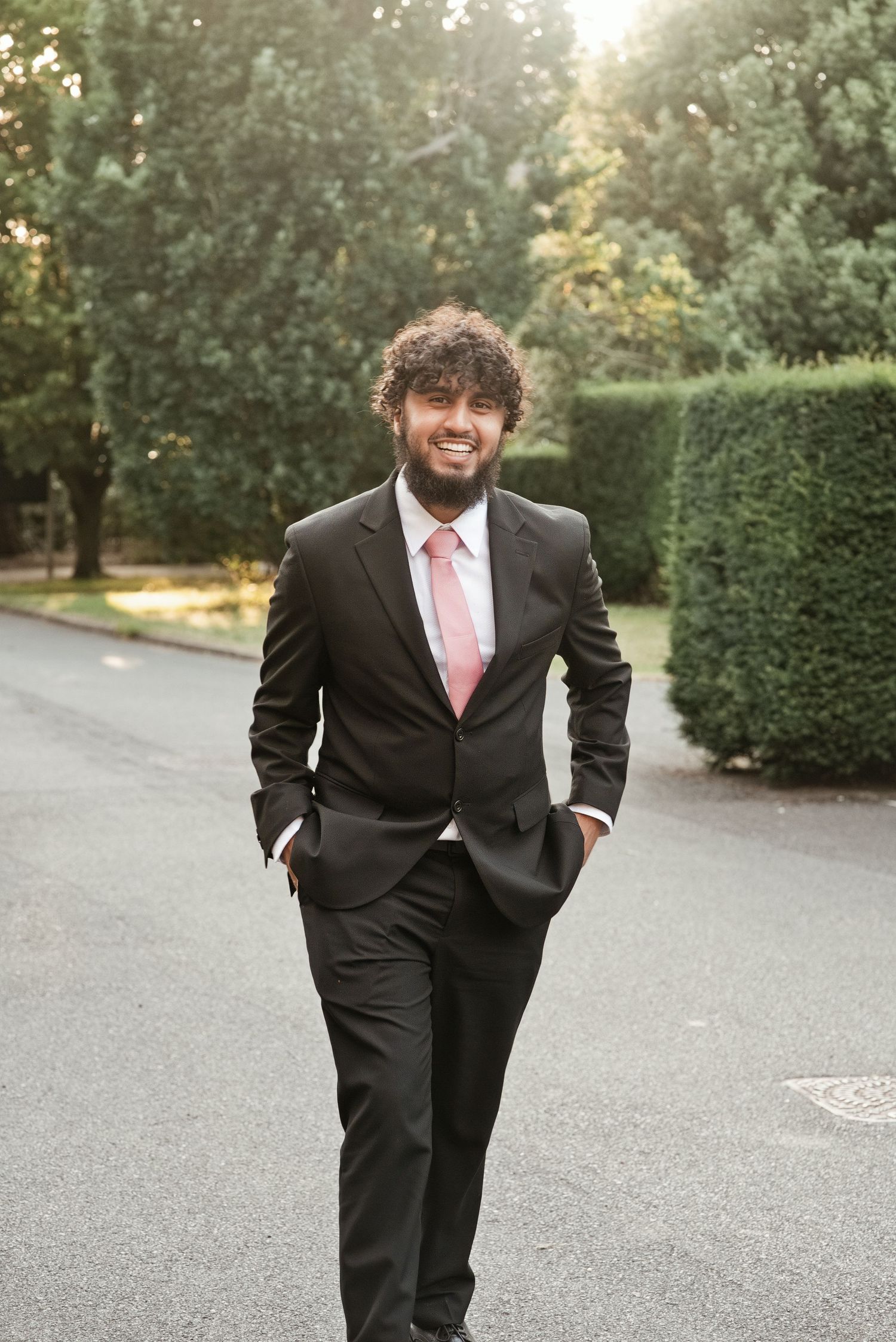 Smiling man in black suit and pink tie walking outdoors in a park setting with green hedges and trees.