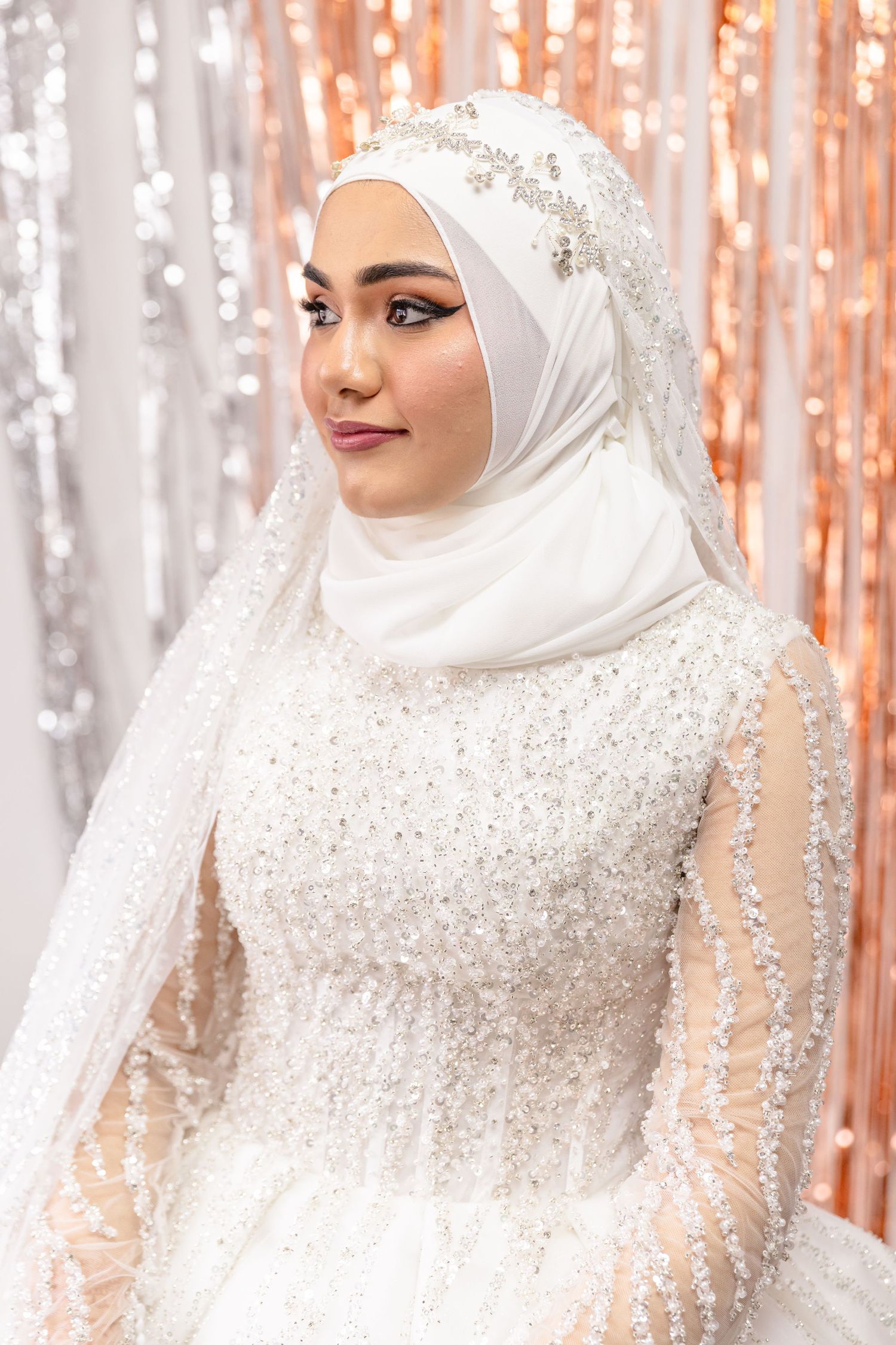 Stunning white wedding dress with intricate beadwork and matching hijab against a shimmering rose gold backdrop.