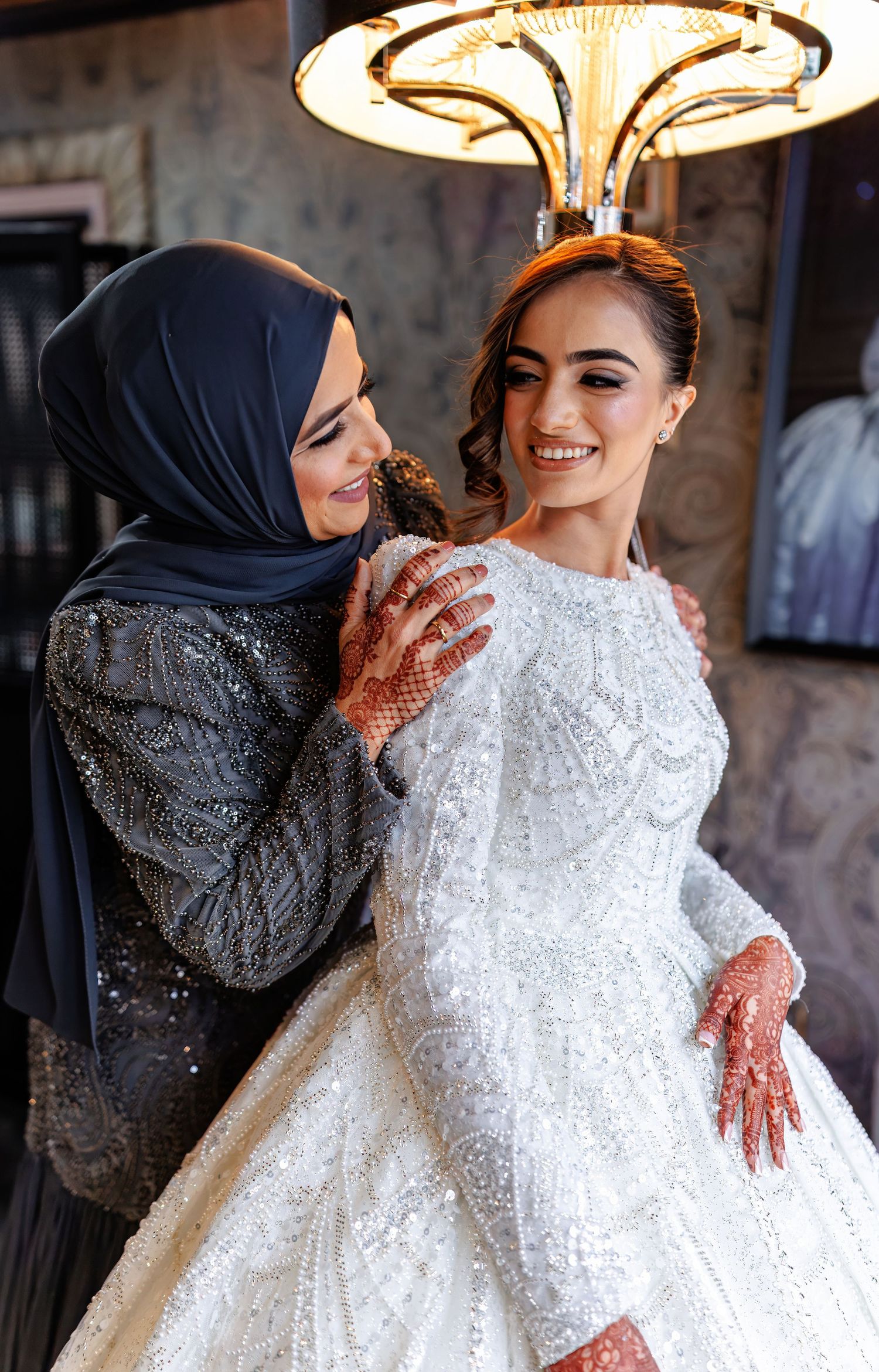 Two women share a joyful moment, one wearing a hijab and sparkly dress, the other in an elegant white wedding gown.