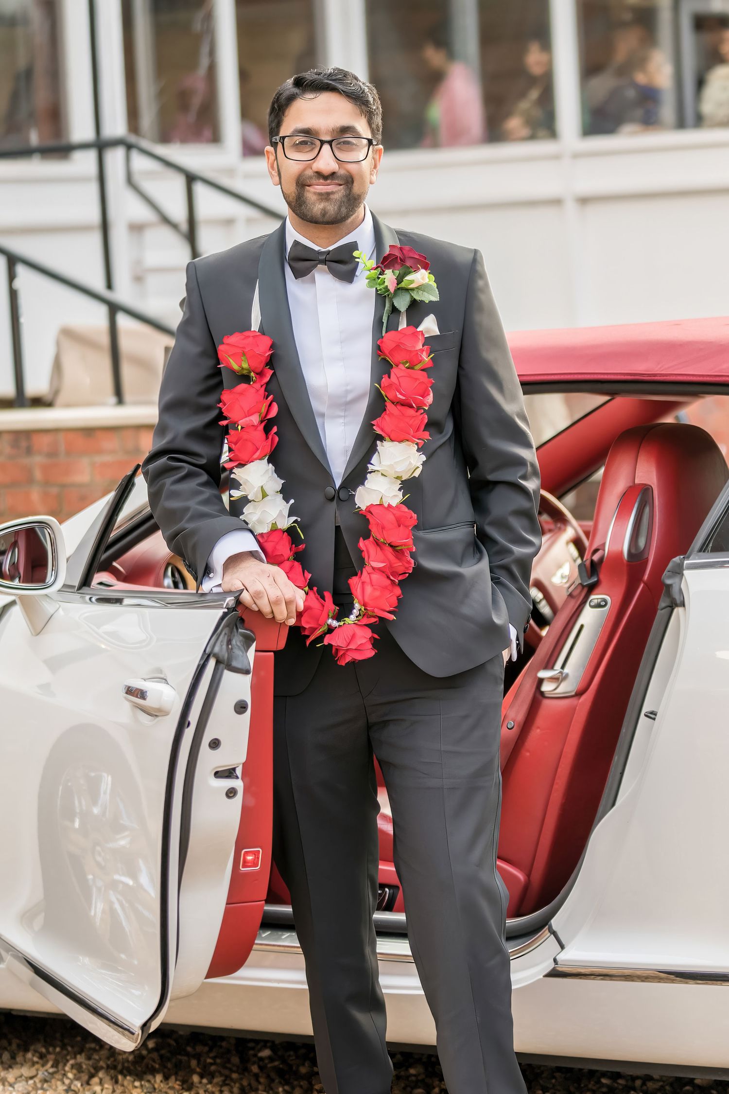 Person in formal tuxedo with red and white flower garland standing beside white luxury car.