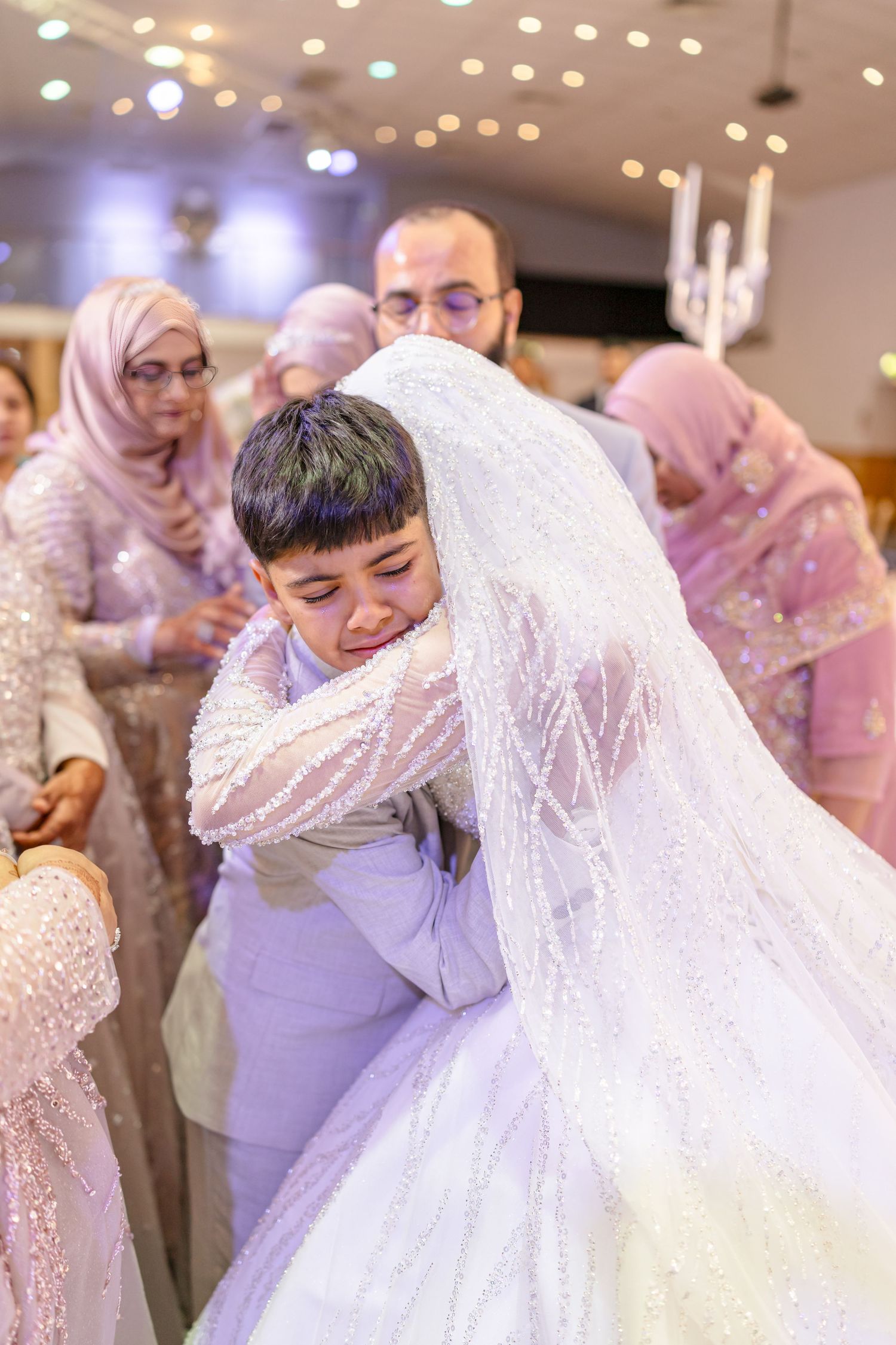 An emotional embrace at a wedding reception with guests in pink and purple attire looking on.