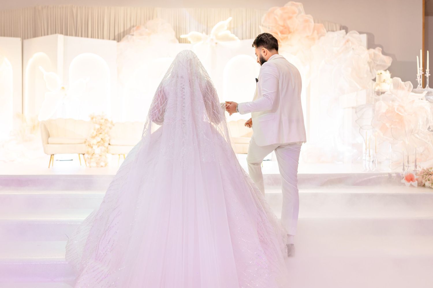 A bride and groom in white attire standing together on a softly lit stage with elegant floral decorations.