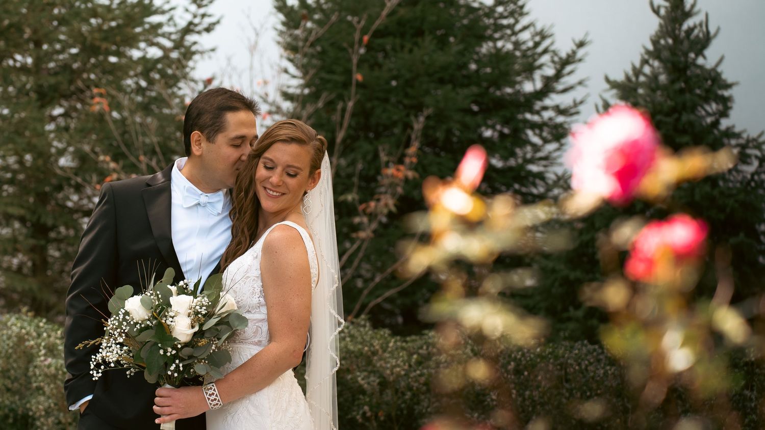 Couple in formal wedding attire sharing intimate moment outdoors with evergreen trees and blurred pink flowers in foreground.