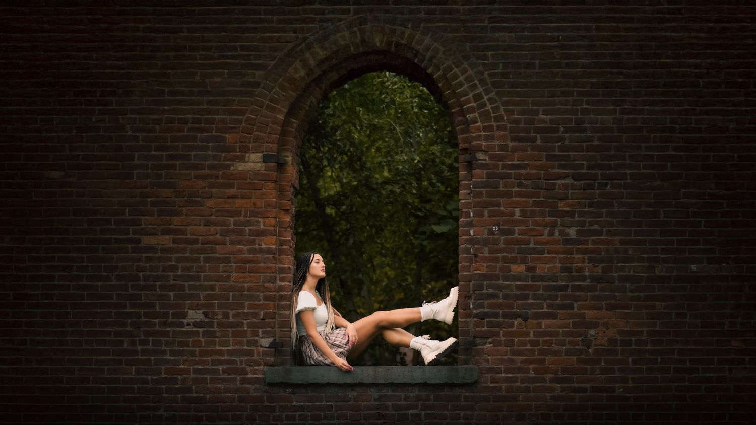 A person rests in a brick archway opening overlooking greenery in an artistic outdoor portrait setting.