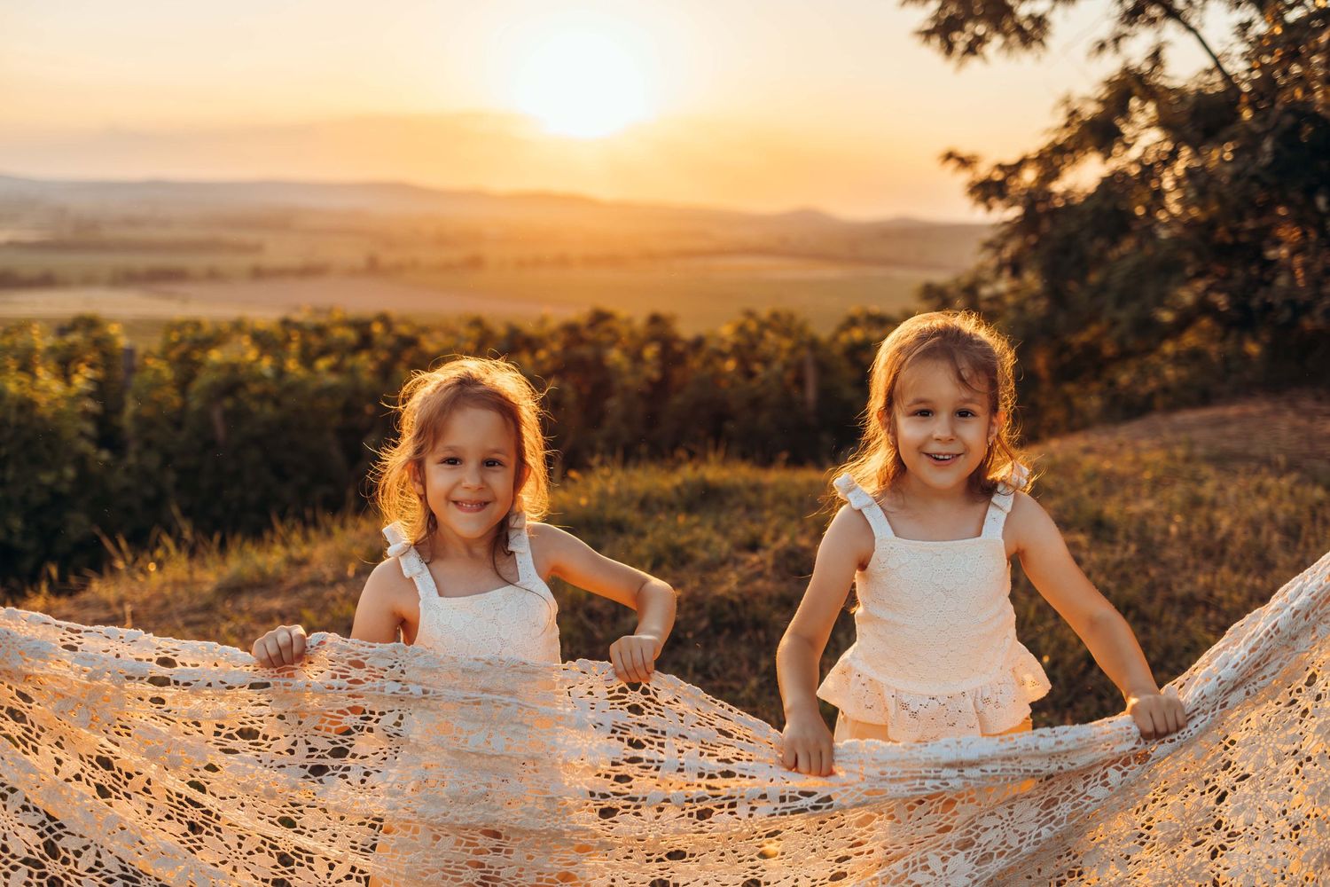 Two children in white dresses pose outdoors at sunset with golden light illuminating rolling hills in the background.