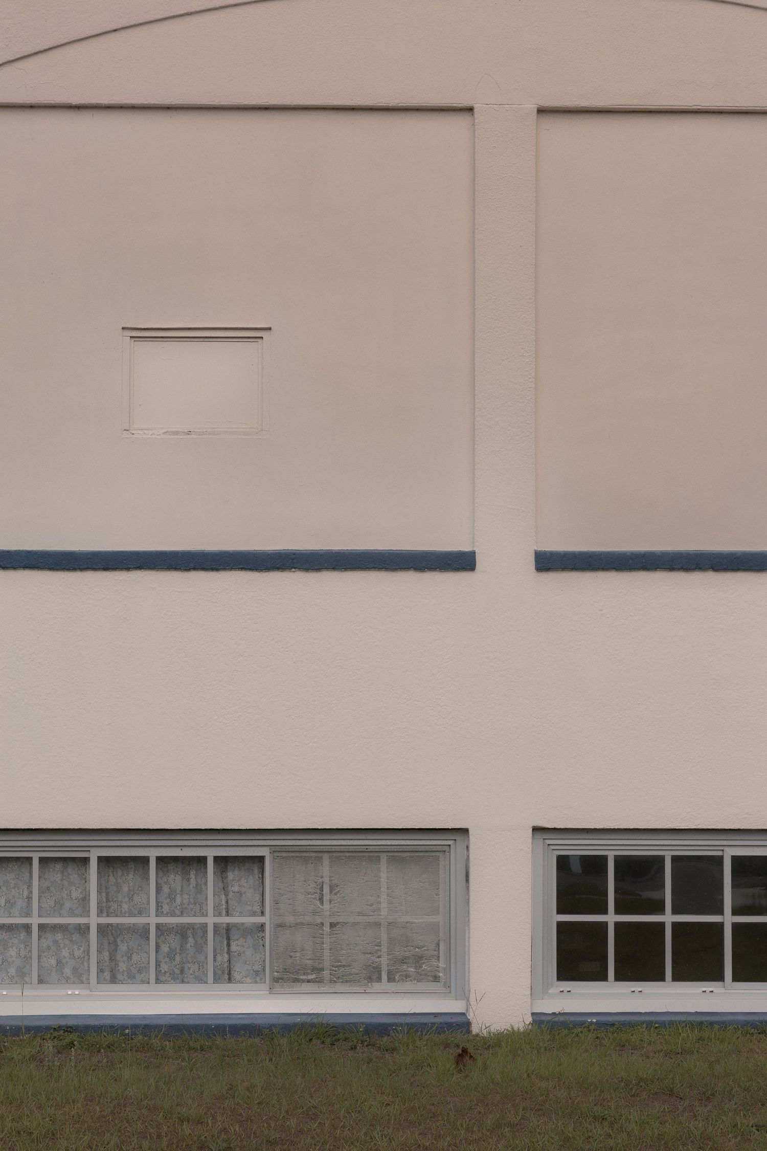 Minimalist exterior wall of a building with large windows, blue trim accent, and small upper window on beige facade.