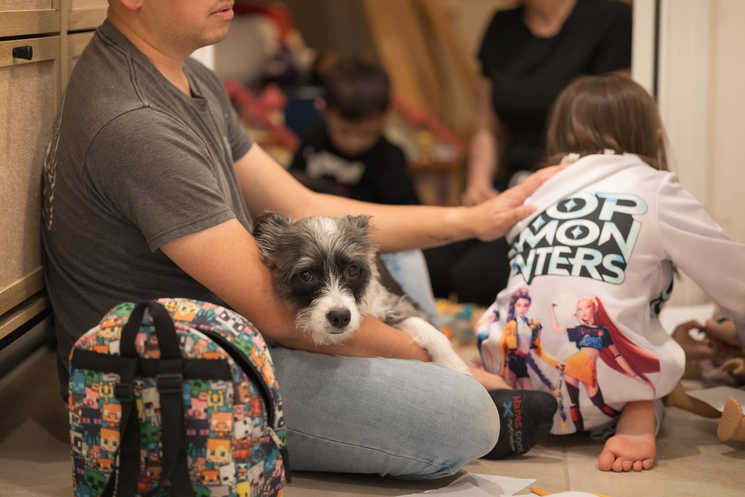 A small scruffy dog sits with people on the floor near a floral backpack.