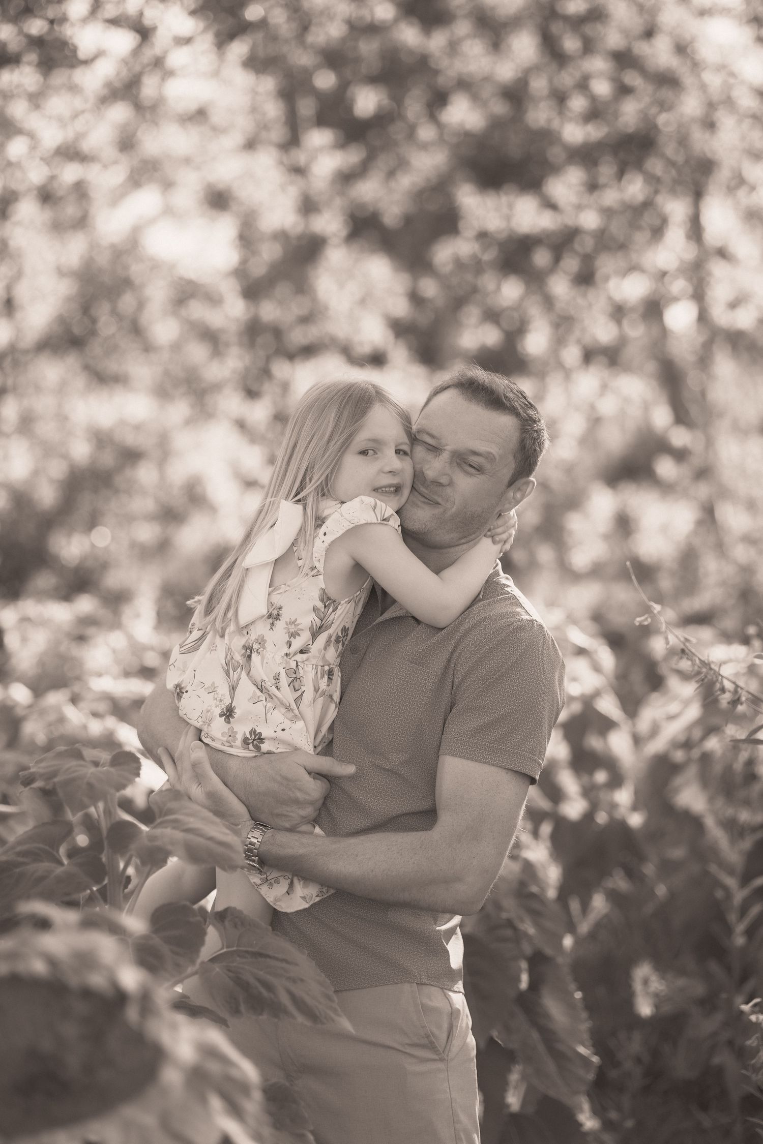 Black and white photo of a father holding his young daughter outdoors, sharing a tender moment surrounded by trees.