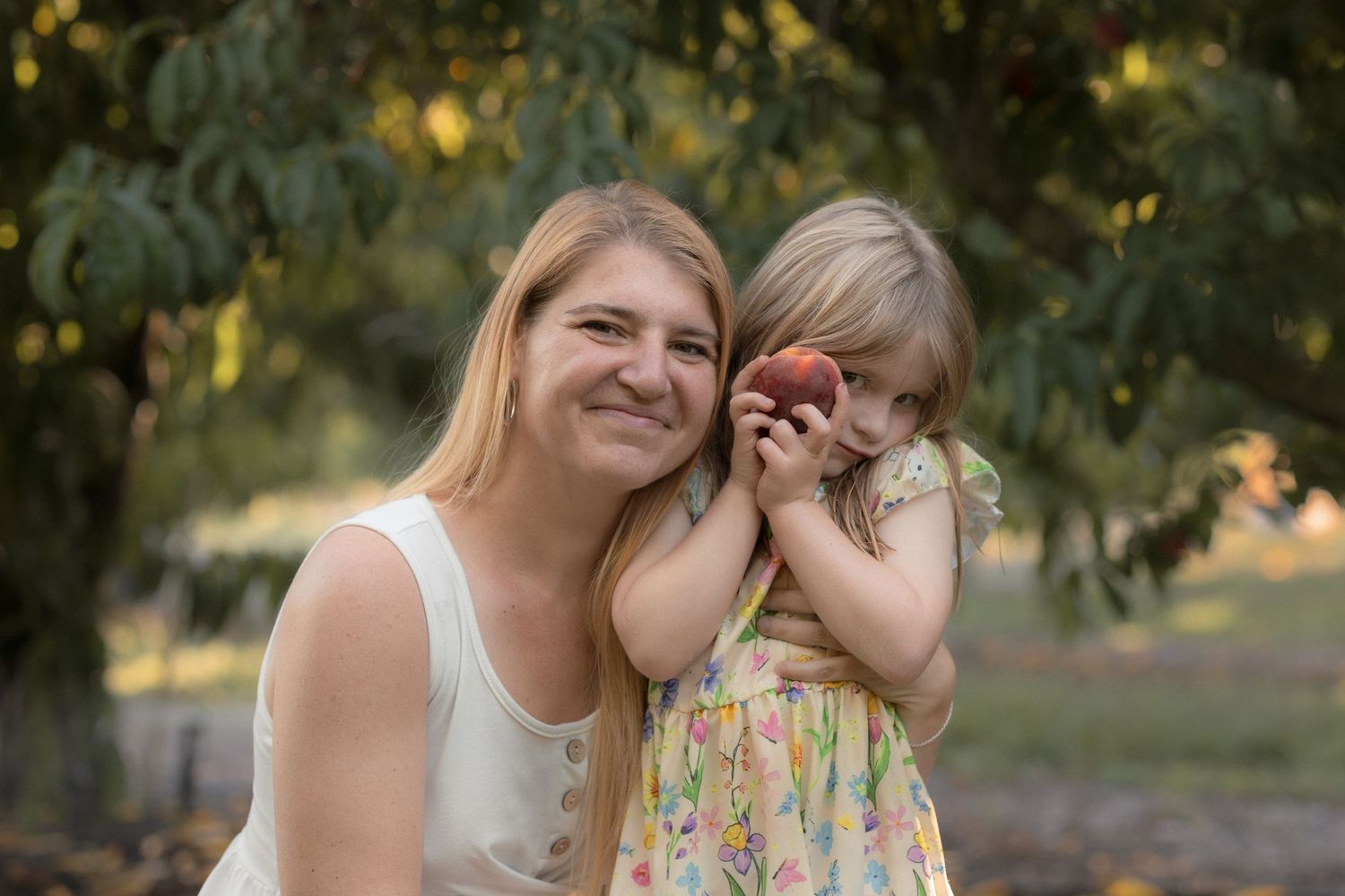 Woman and young girl holding a peach together in an orchard, smiling outdoors among fruit trees.