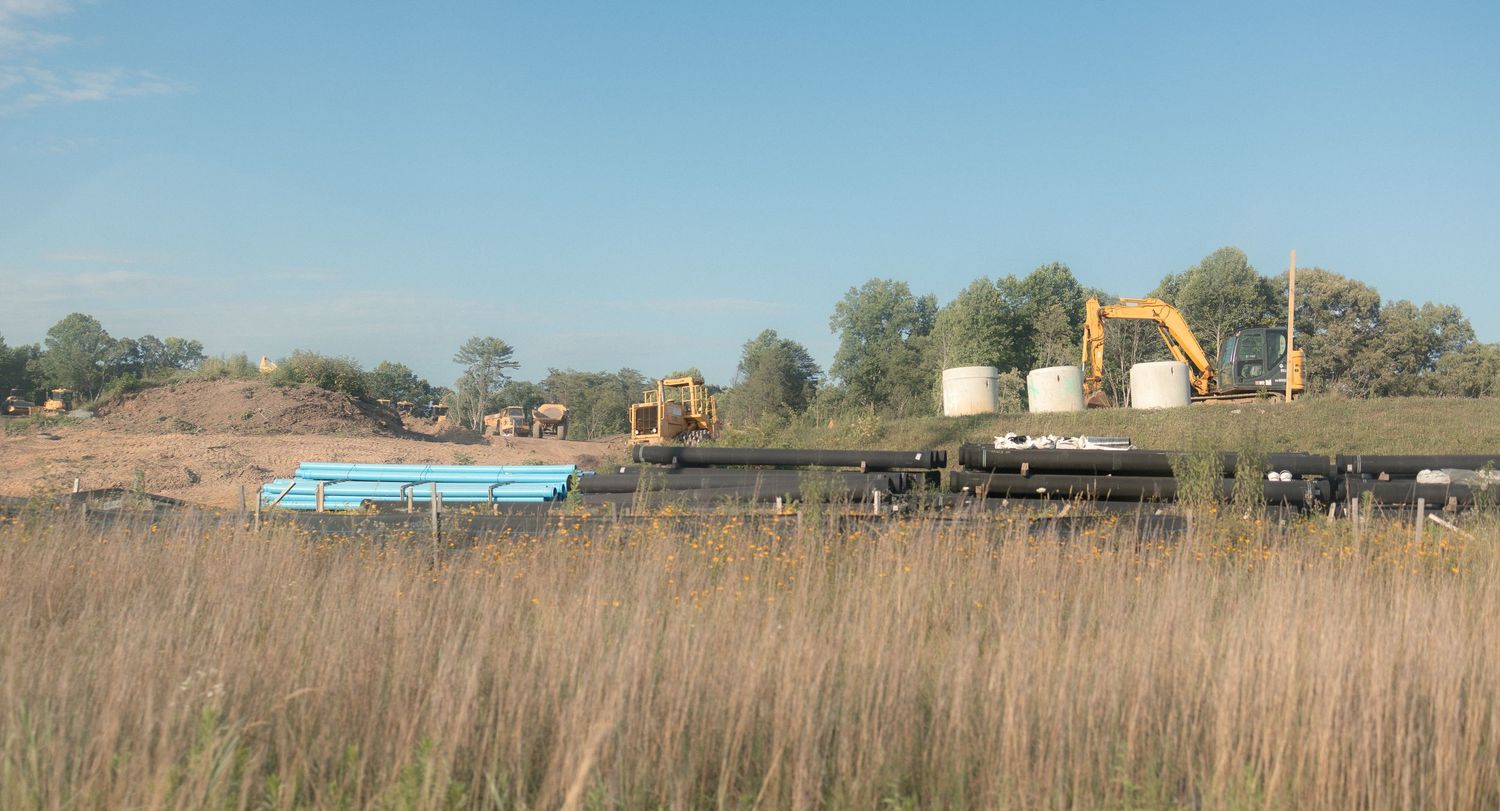 Construction site with excavators, storage tanks, and blue pipes amid flat grasslands under a clear sky.