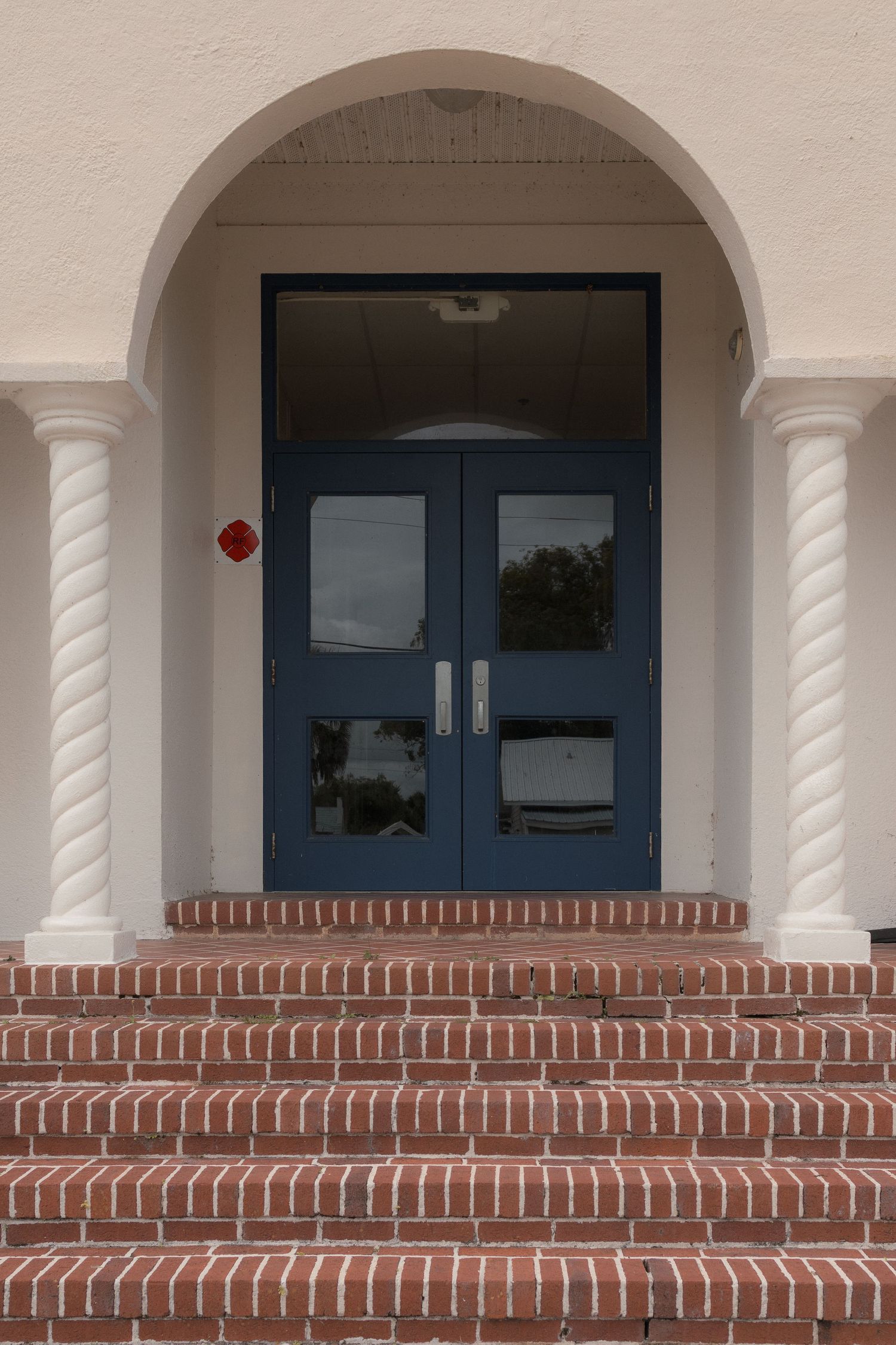 Blue double doors with glass panels framed by a white arch and spiral columns, atop red brick steps.