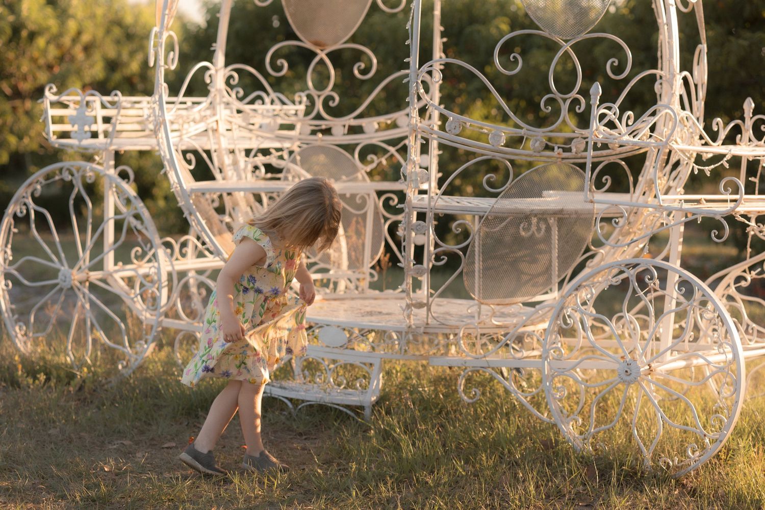 A young girl in a floral dress plays beside a white ornate fairy tale carriage prop outdoors at golden hour.