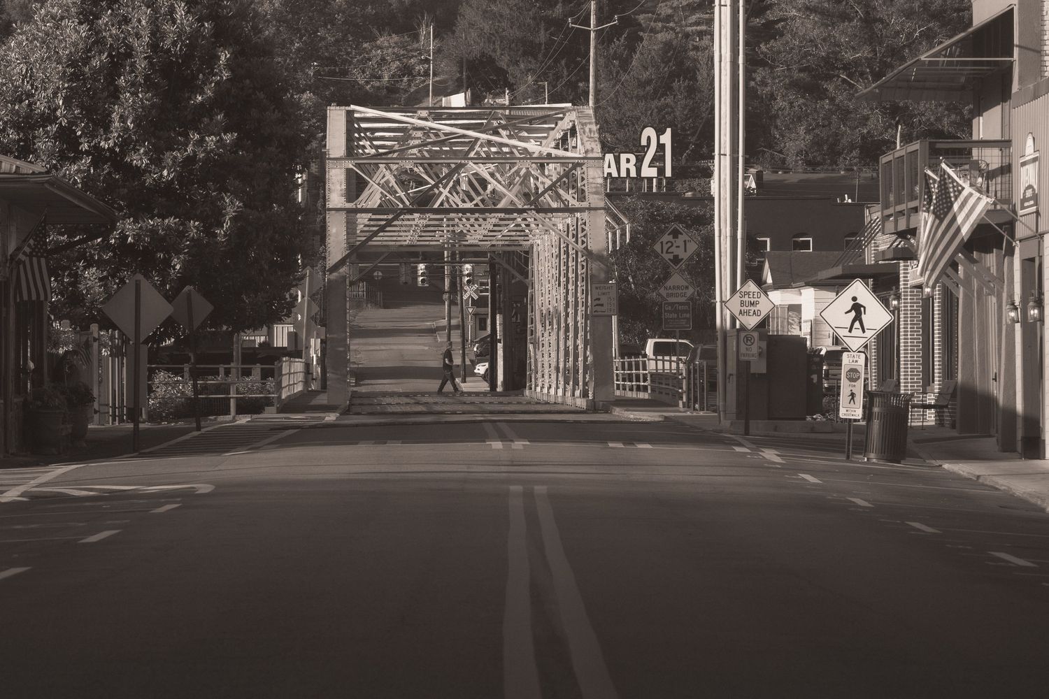 Black and white photo of empty street leading to ornate gate with AR 21 sign, trees and traffic signs visible.