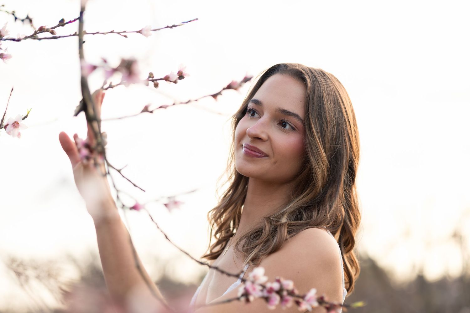 Woman in white dress poses gracefully among blooming cherry blossom branches during golden hour sunset.