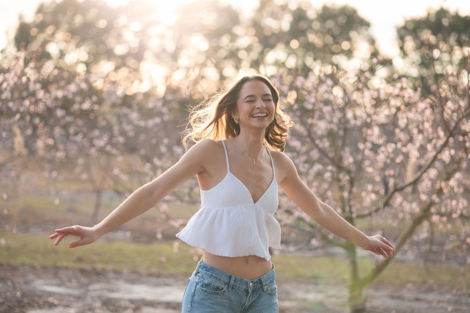 Dancing in a blooming orchard at sunset wearing a white crop top and jeans with golden light filtering through trees.