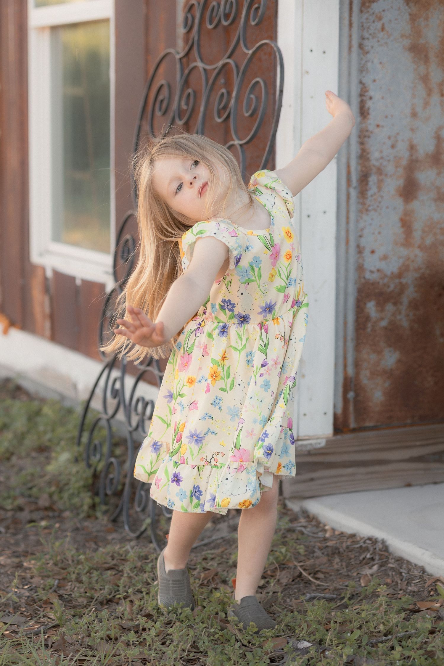 Young girl in floral dress twirling outdoors near rustic building, long blonde hair flowing, arms outstretched joyfully.