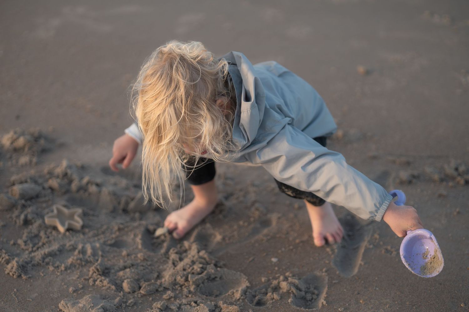Person in blue jacket and light pants crouches down to explore sandy beach at dusk.