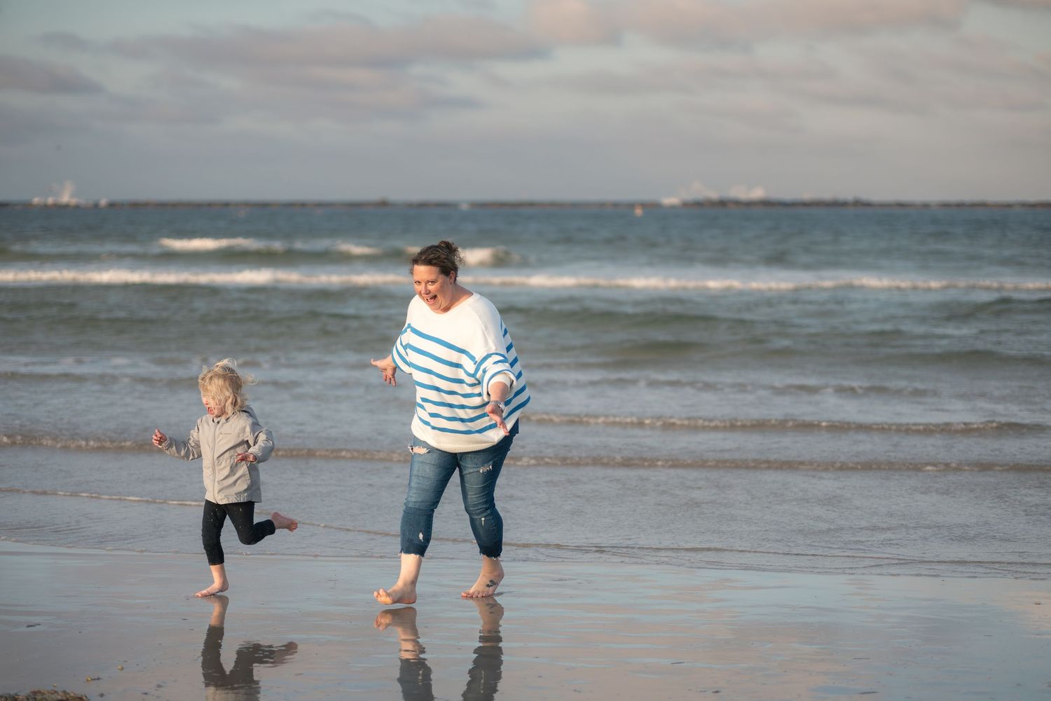 Two people run and play together along a sandy beach at sunset with ocean waves in the background.