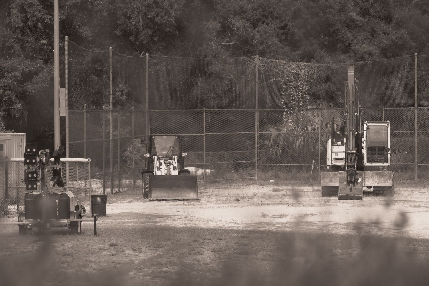 Black and white photo of construction equipment including excavators near a chain-link fence at an outdoor work site.