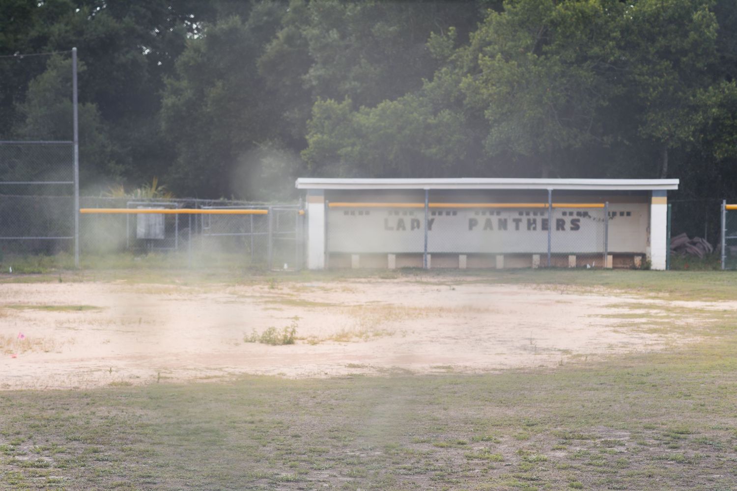 Dusty Lady Panthers softball field with dugout and lush green trees in background on a hazy day.