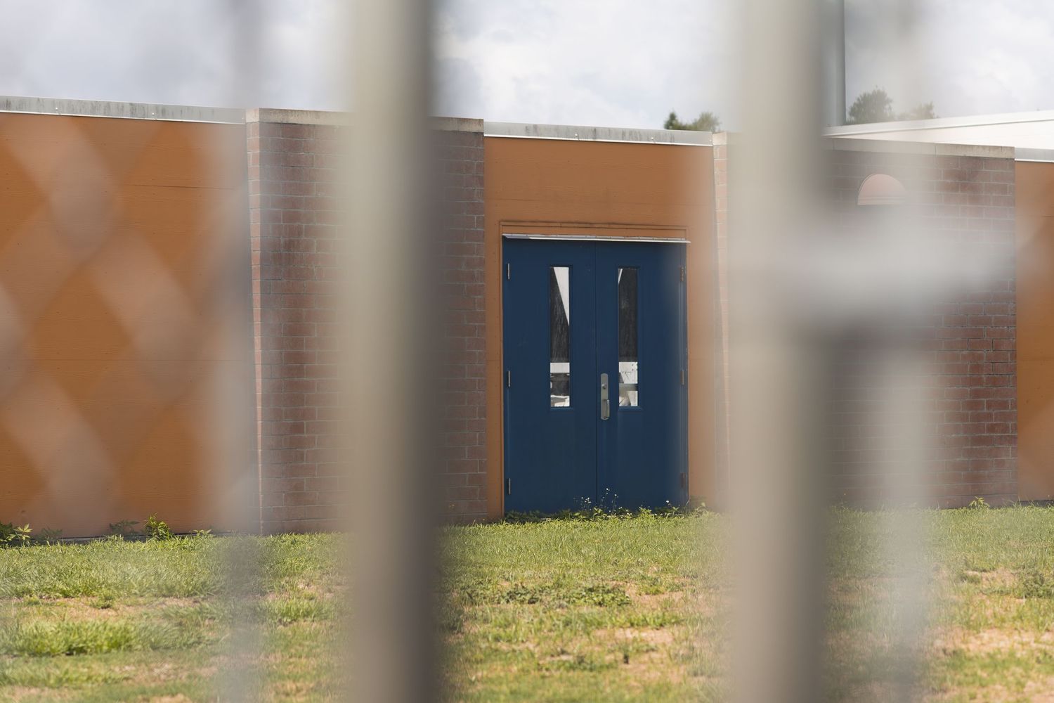 Blue double doors on a brick building viewed through a chain-link fence at a secure facility.