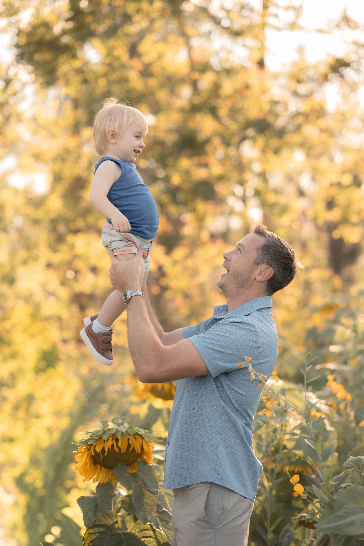 A father joyfully lifts his smiling toddler in a sunflower field with golden autumn trees in the background.