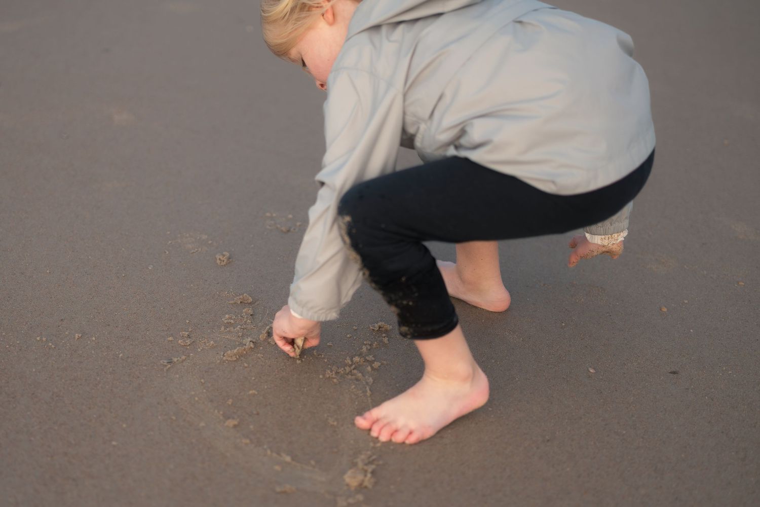 Barefoot person crouches down to examine something in the wet sand on a beach.